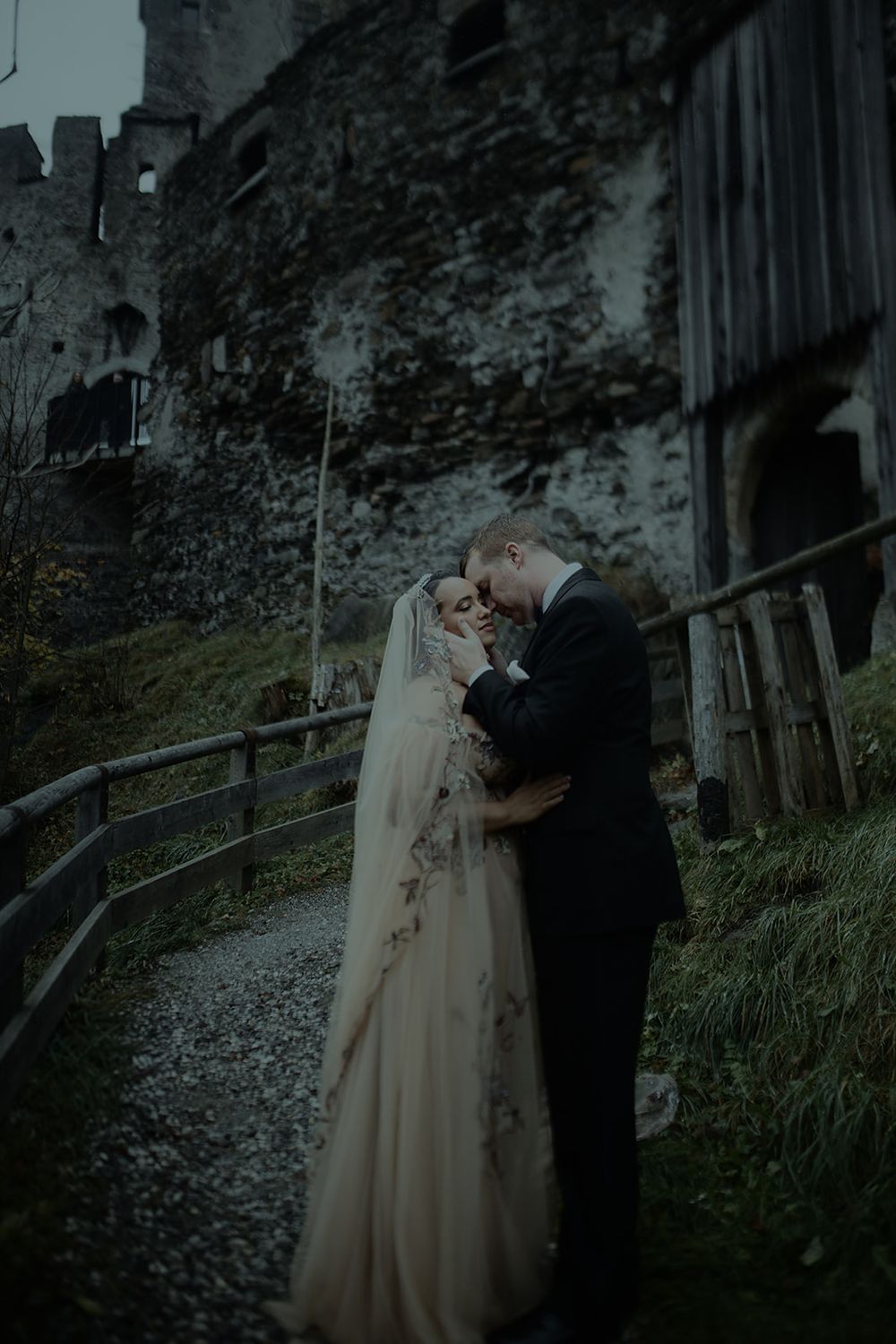 Bride and groom embrace romantically before a dark medieval castle on their wedding day.