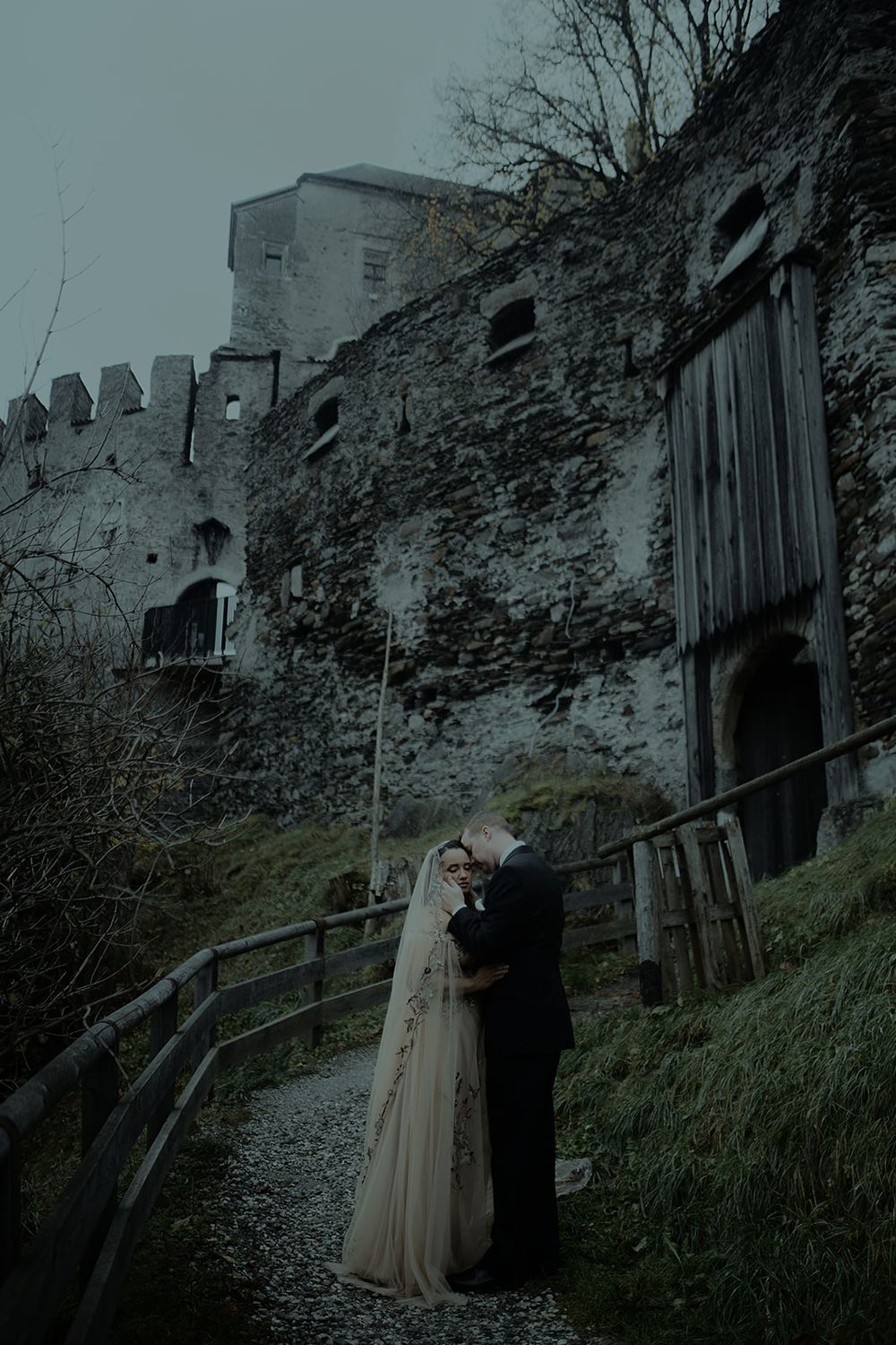 Bride and groom embracing on path before ancient castle ruins in moody atmospheric setting.