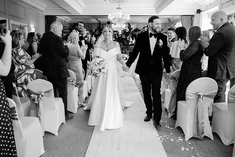 Black and white photo of a bride and groom walking down the aisle at their elegant wedding ceremony.