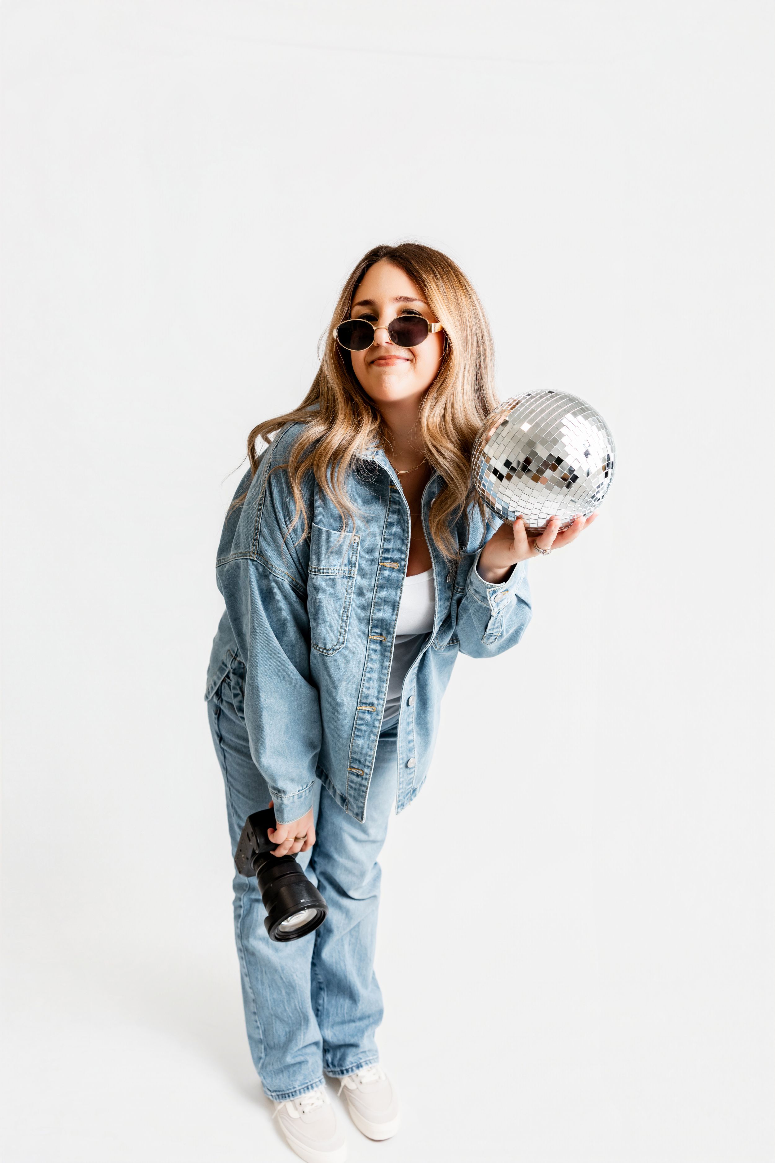 Woman in denim outfit holding disco ball and camera, wearing round sunglasses, posing on white background.