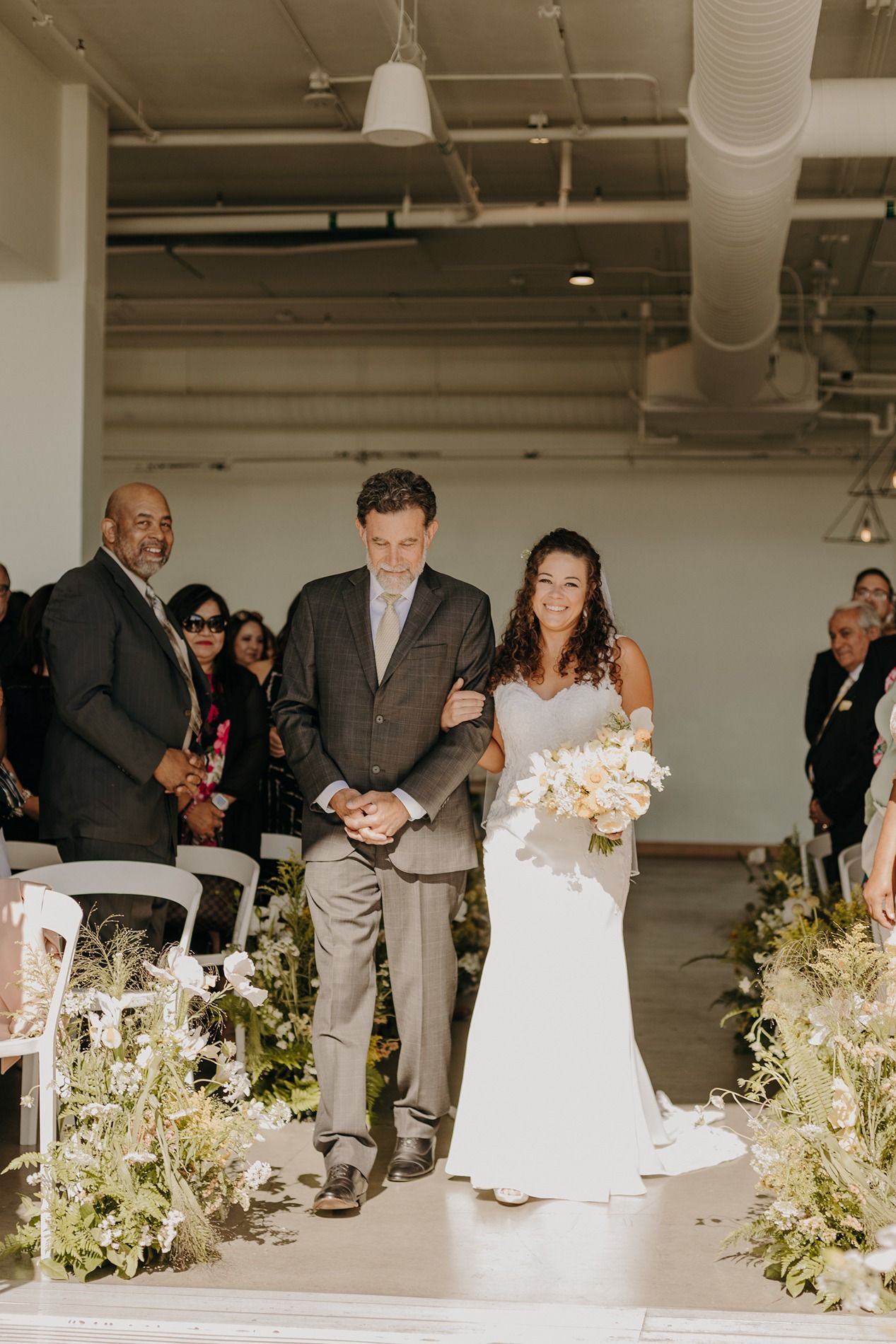 Bride in white gown holding bouquet walks down aisle with escort at indoor wedding ceremony at The Lane.