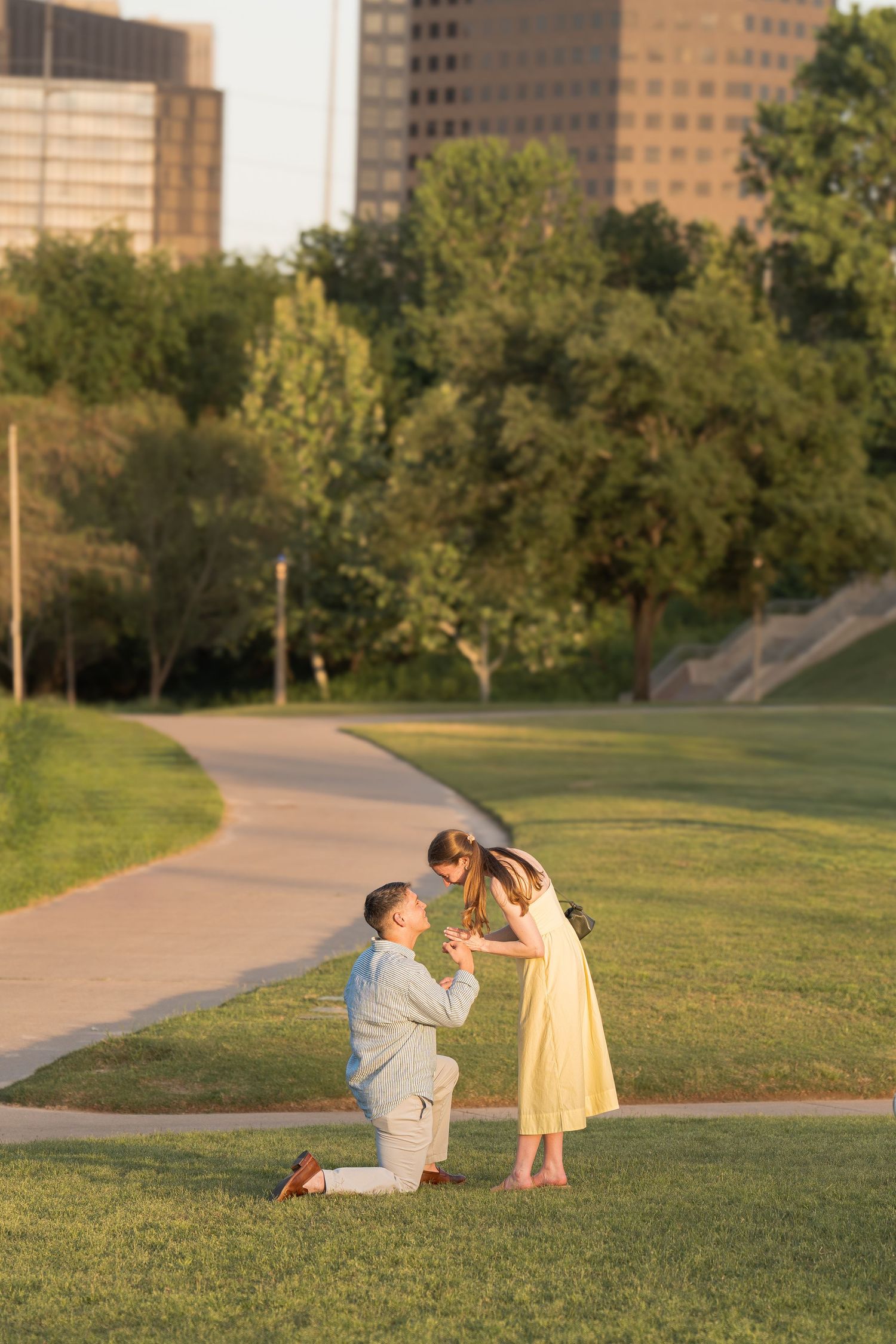 Man kneeling on one knee proposing to woman in yellow dress in a sunny park with city buildings in background.