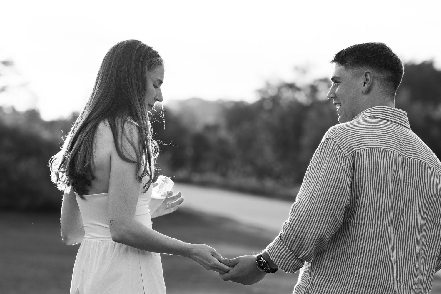 Black and white photo of a couple holding hands and smiling at each other outdoors.