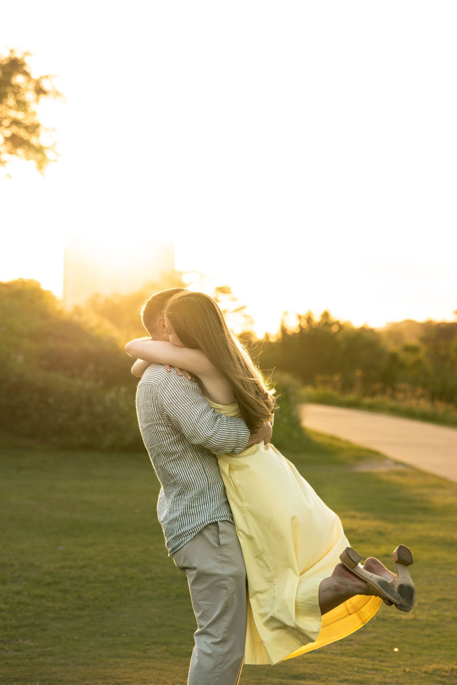 Couple sharing a romantic embrace at sunset in a lush green park, woman in yellow dress lifted off ground.