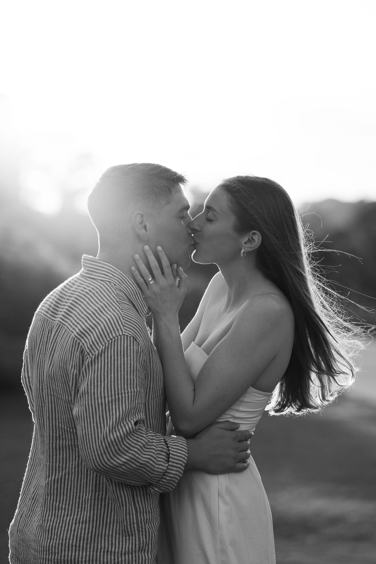 Black and white photo of a romantic couple sharing a kiss outdoors, with wind blowing the woman's long hair.