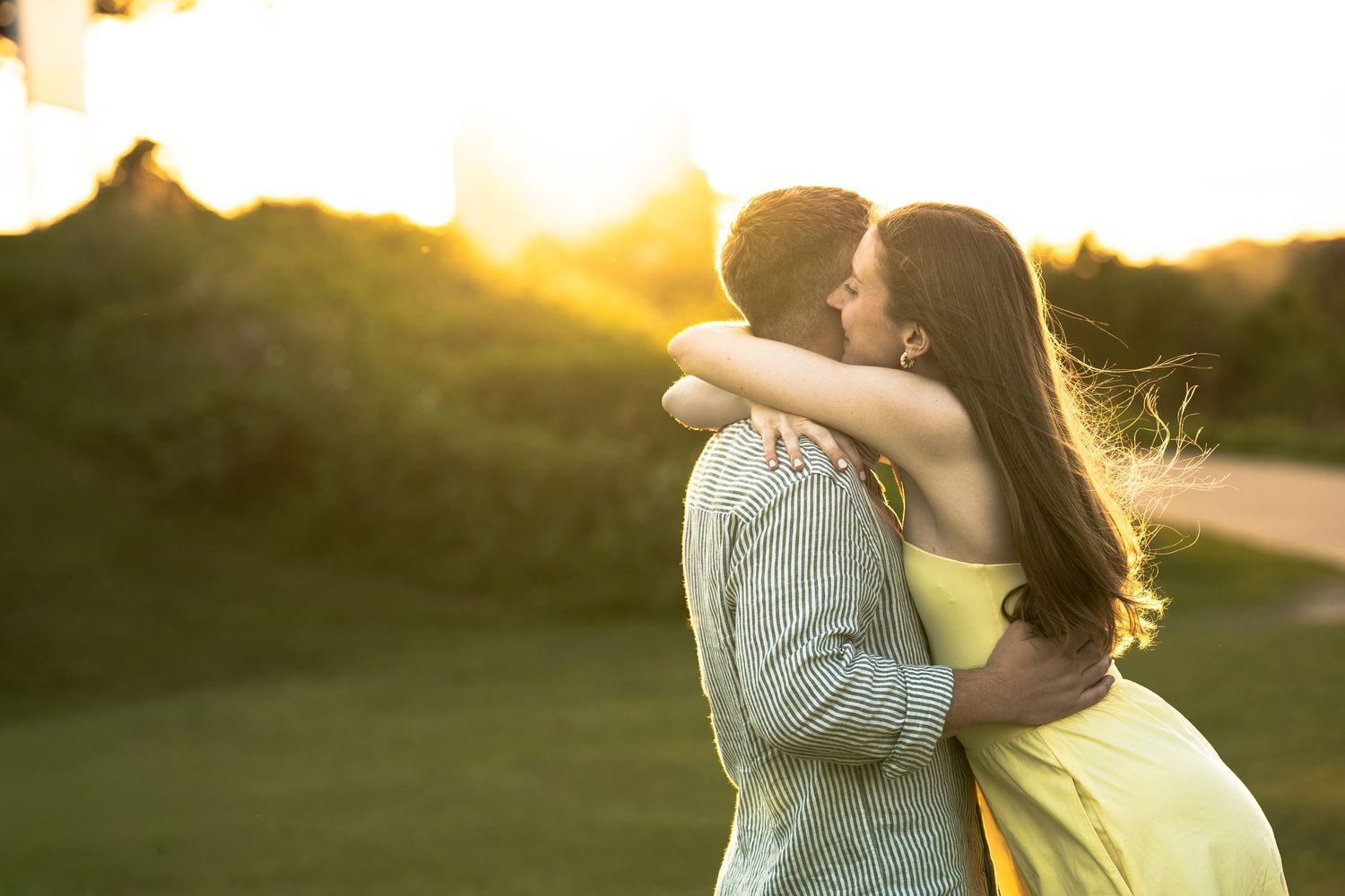 Couple sharing a romantic kiss at golden sunset, woman in yellow dress with long dark hair outdoors.