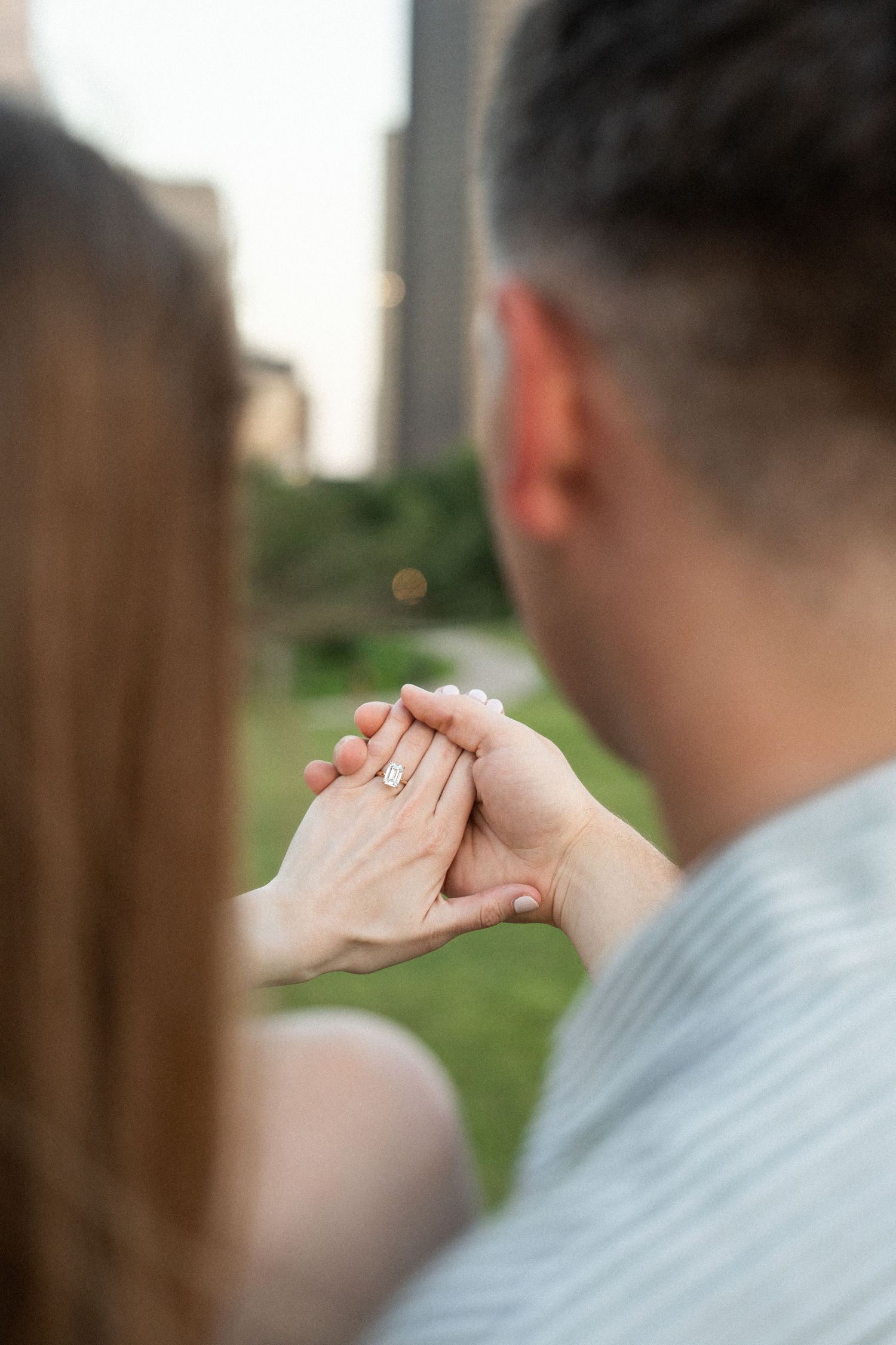 Couple holding hands displaying a diamond engagement ring in an urban park setting.