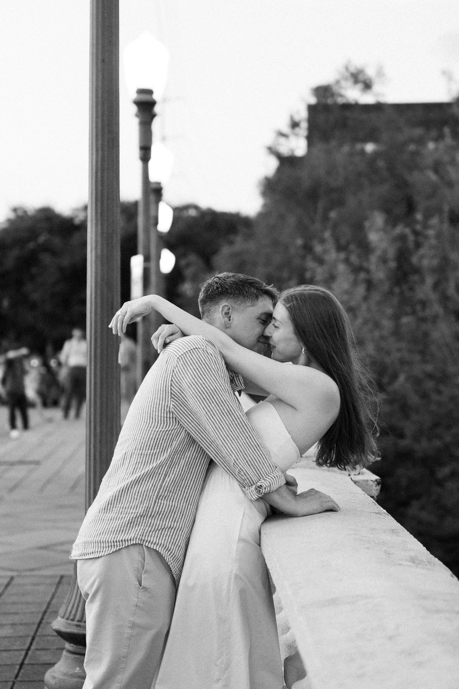 Black and white photo of a romantic couple nearly kissing while embracing near a stone railing outdoors.