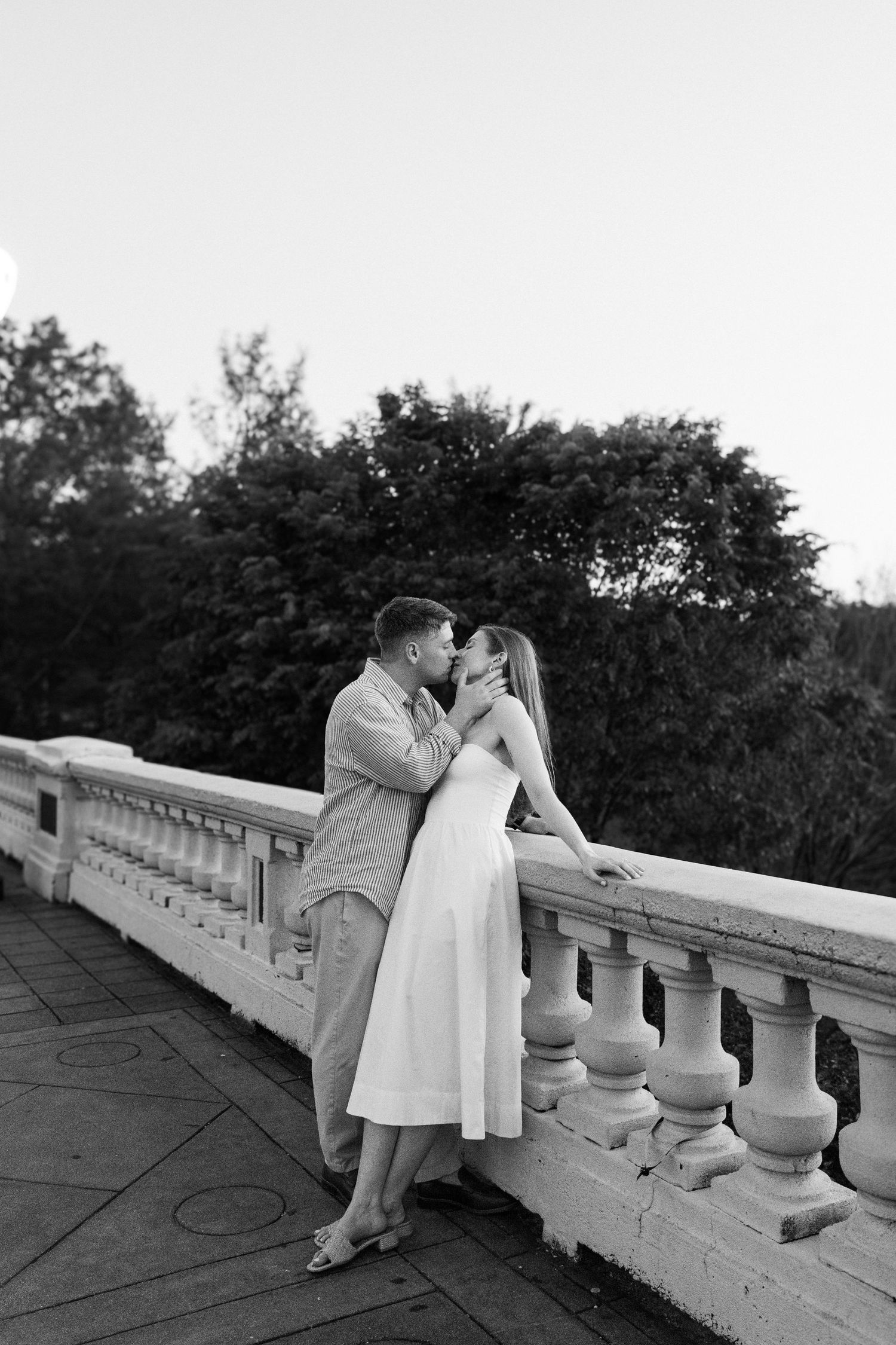 Black and white photo of an engaged couple kissing on a stone balcony surrounded by trees.