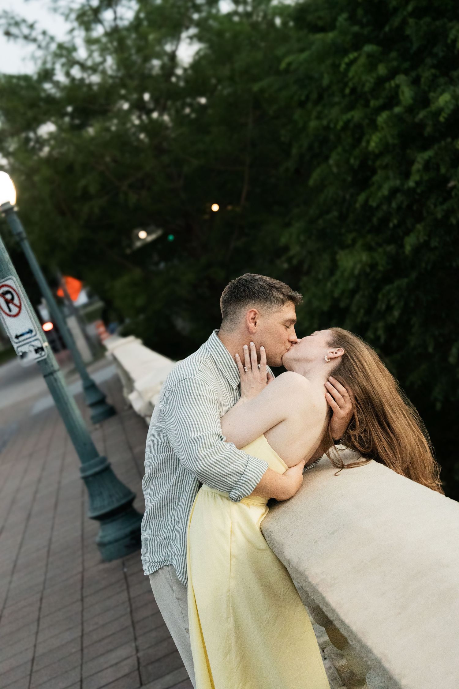 A couple shares a romantic kiss leaning against a stone bridge railing at dusk on a tree-lined street.