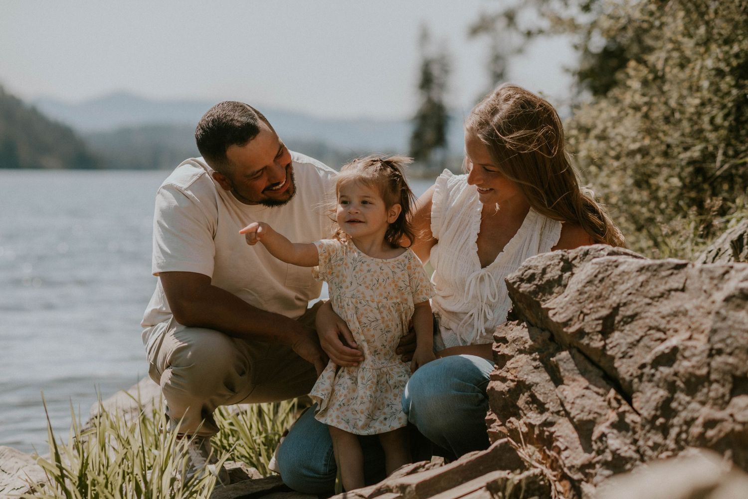 A happy family of three enjoys a sunny day by a mountain lake, with parents smiling at their toddler daughter.