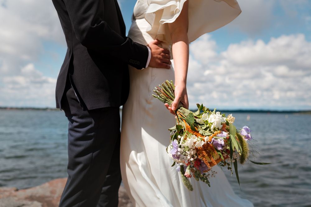 Bride and groom standing by the water, bride holding a colorful wildflower bouquet in a white wedding dress.