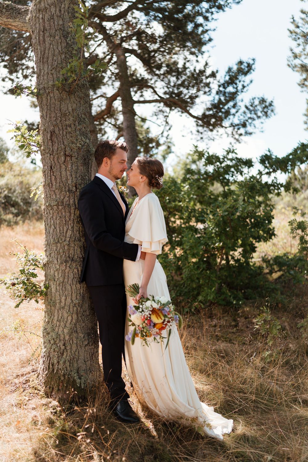 Bride and groom share a romantic moment leaning against a tree in a sunlit outdoor setting, bride holding a colorful bouquet.