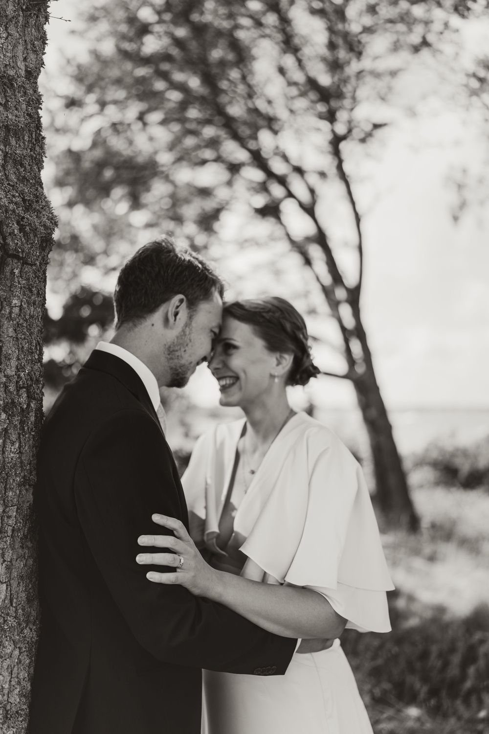 Black and white photo of a smiling bride in white dress embracing groom in suit near a tree outdoors.
