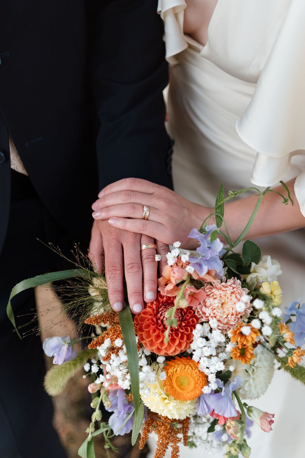 Newlyweds displaying wedding rings on hands resting over a colorful bridal bouquet with orange dahlias and wildflowers.