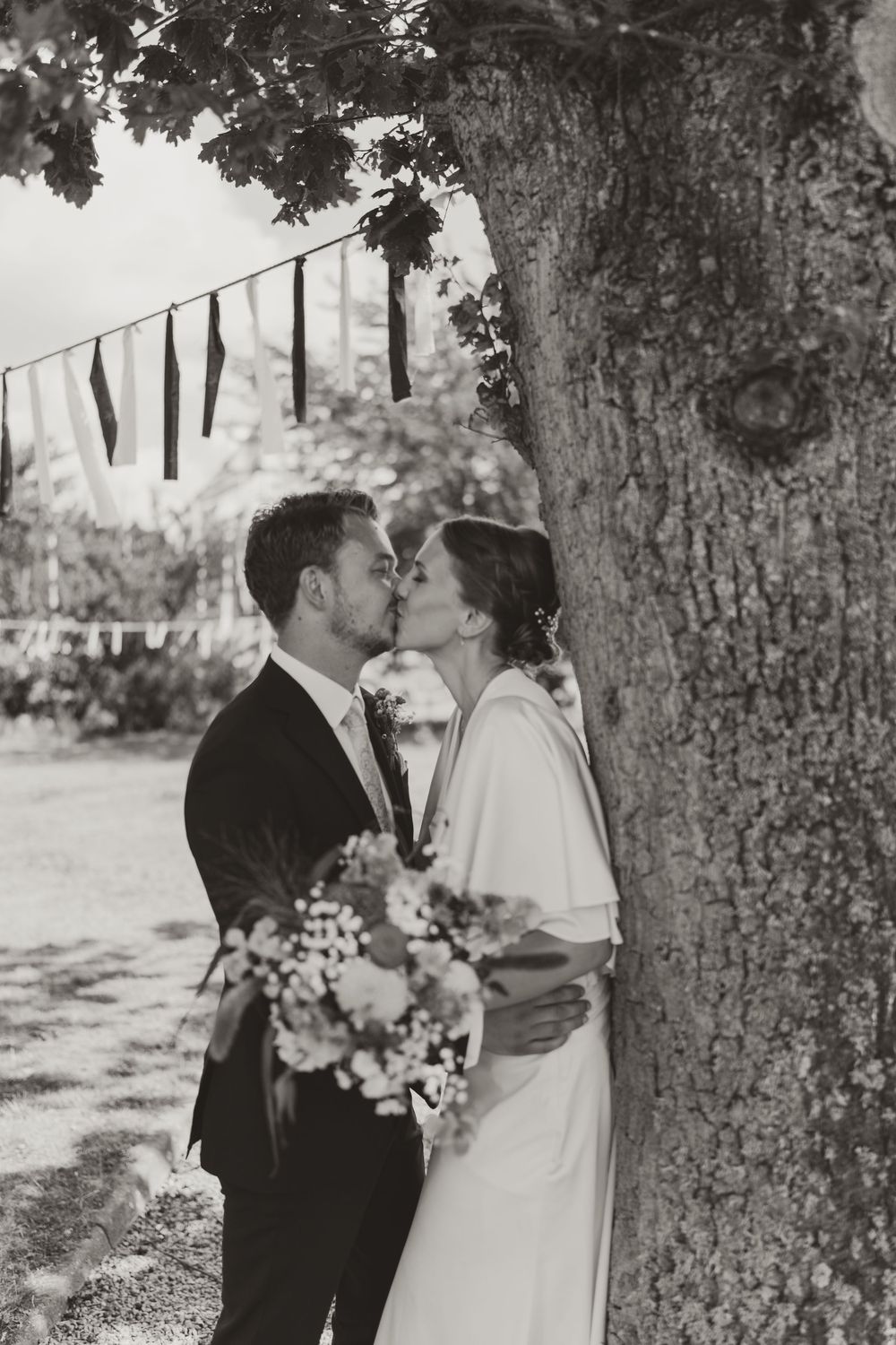Black and white photo of a bride and groom sharing a kiss beside a large tree, bride holding a floral bouquet.