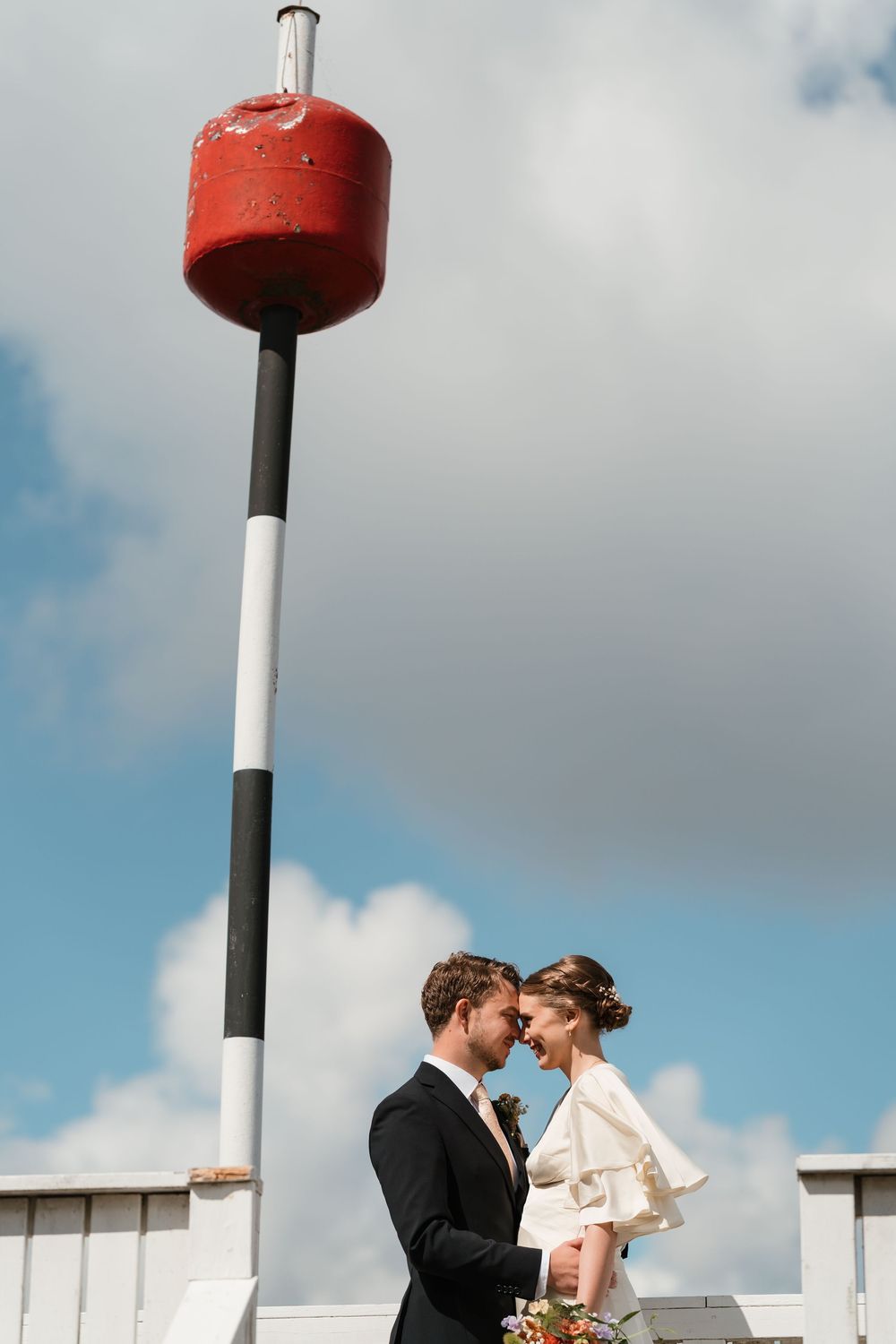 Bride and groom share a romantic moment under a red maritime buoy against a blue sky.