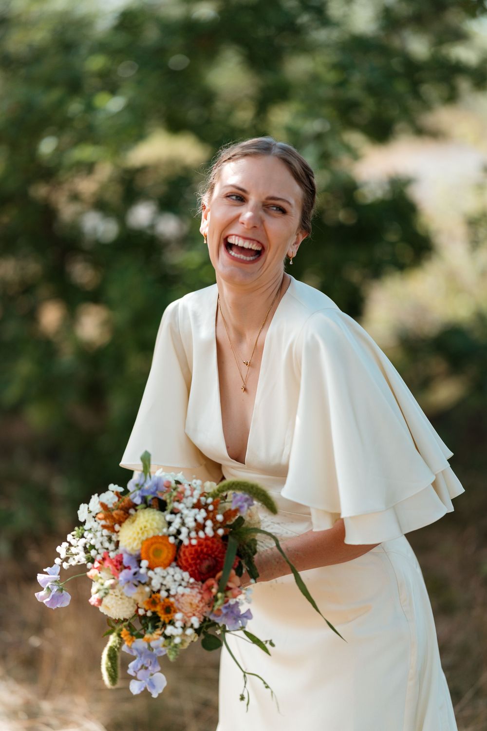 Laughing bride in white cape dress holding colorful wedding bouquet outdoors.