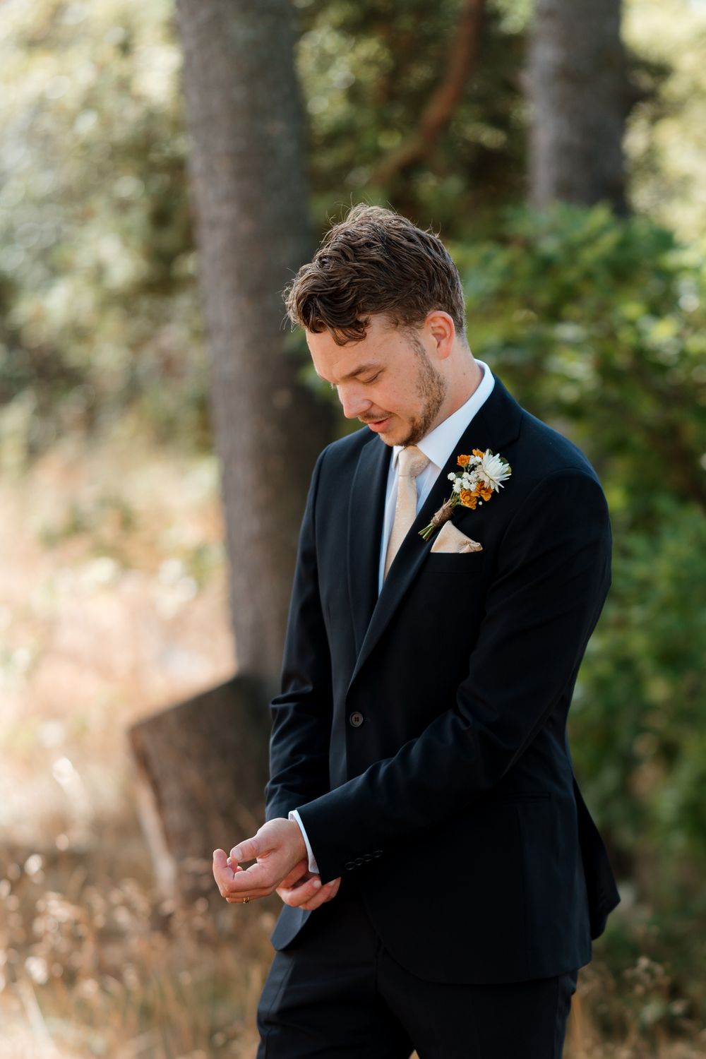 Groom in black suit with floral boutonniere adjusting cufflinks outdoors among trees on wedding day.