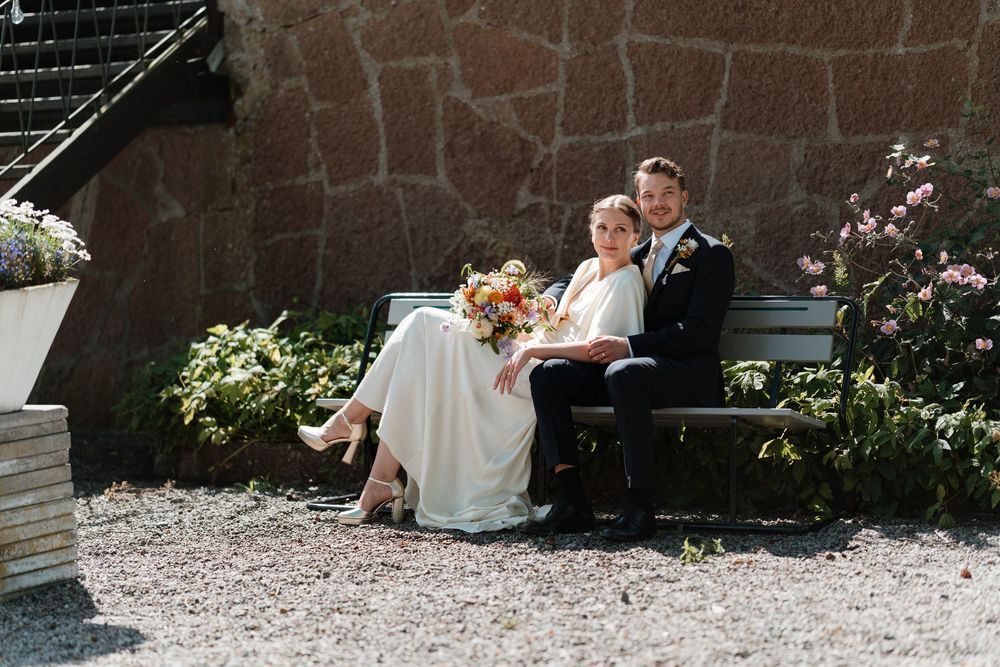 Bride and groom sitting together on a bench outdoors, bride holding a colorful bouquet, against a stone wall backdrop.