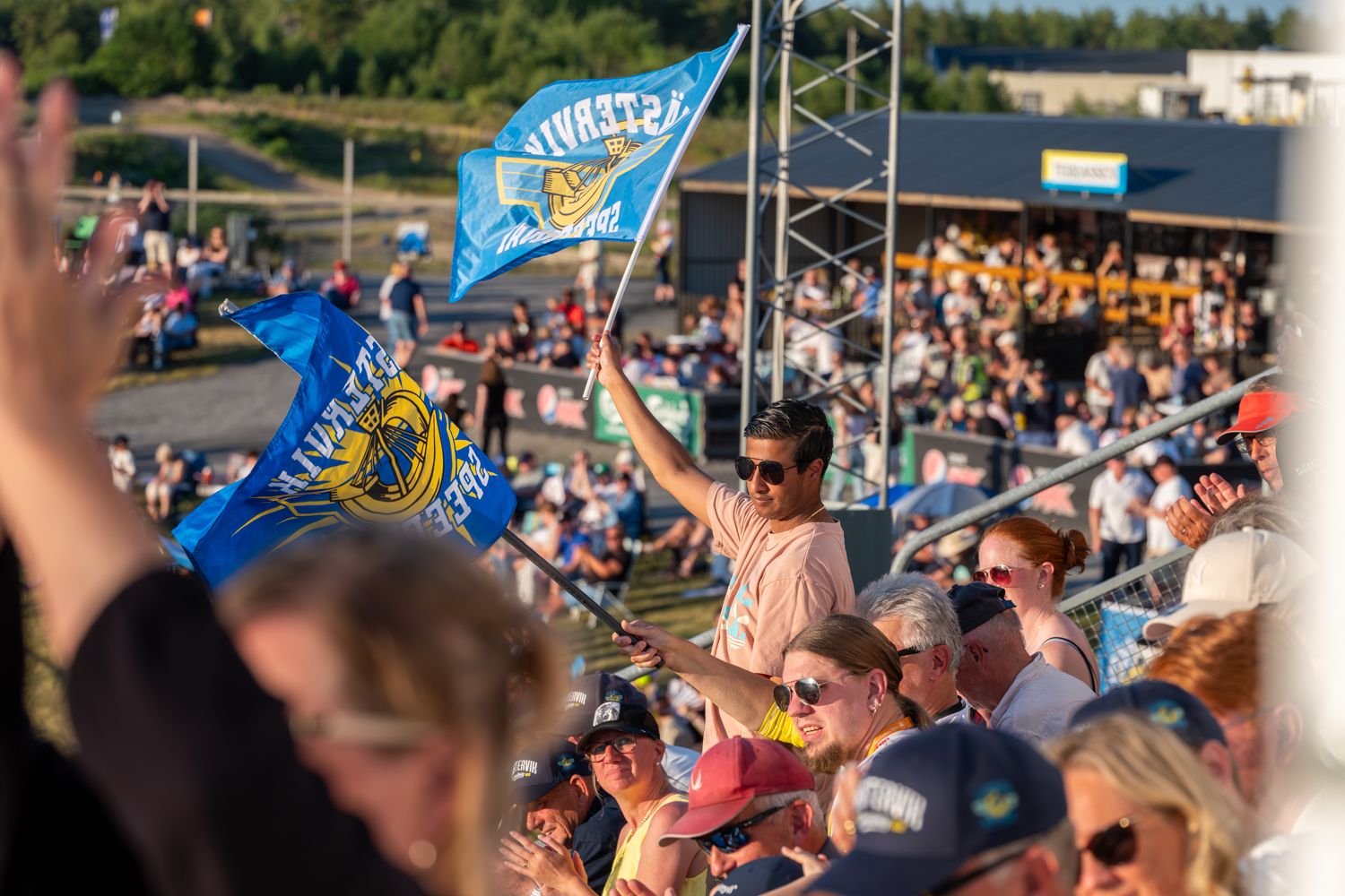 En person står mitt i folkhavet på en läktare och viftar med Västervik Speedways flaggor under en speedwaymatch en varm sommarkväll.