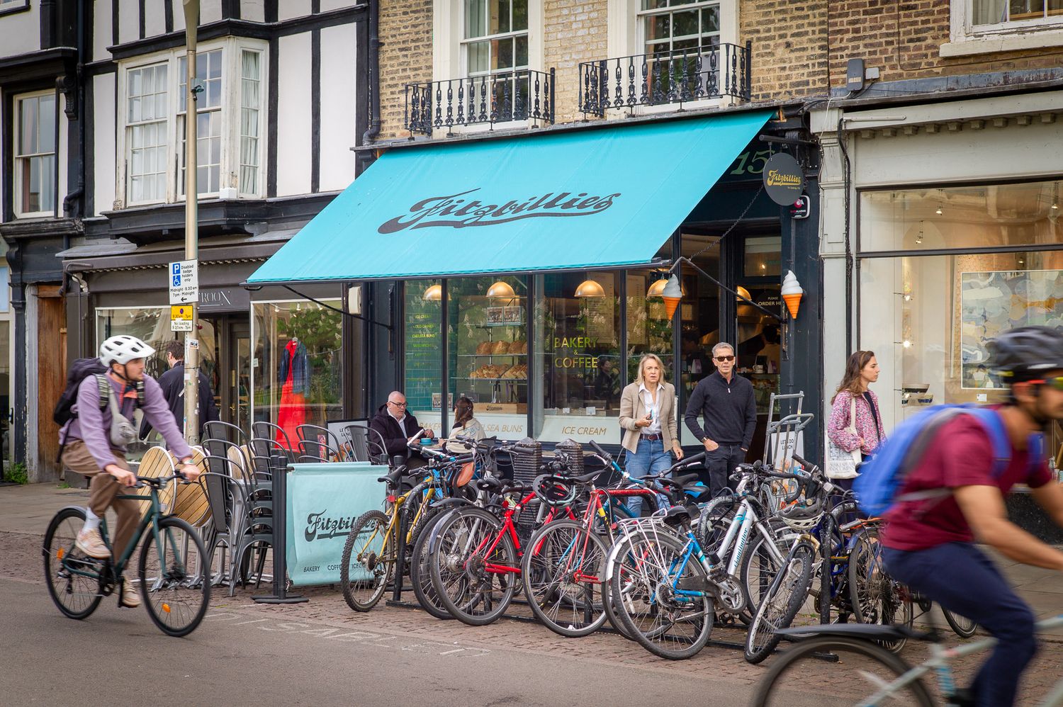 Cyclists and pedestrians outside Fitzbillies cafe with teal awning on a busy Cambridge street.
