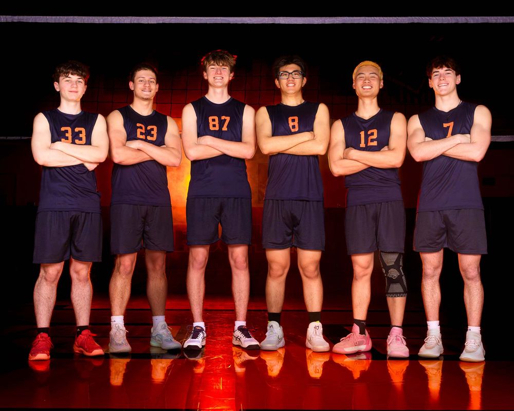 Six Hershey Trojans smile with arms crossed in front of a orang-lit volleyball net by Deanna Fox, SportsFox Photography based in Hershey PA.