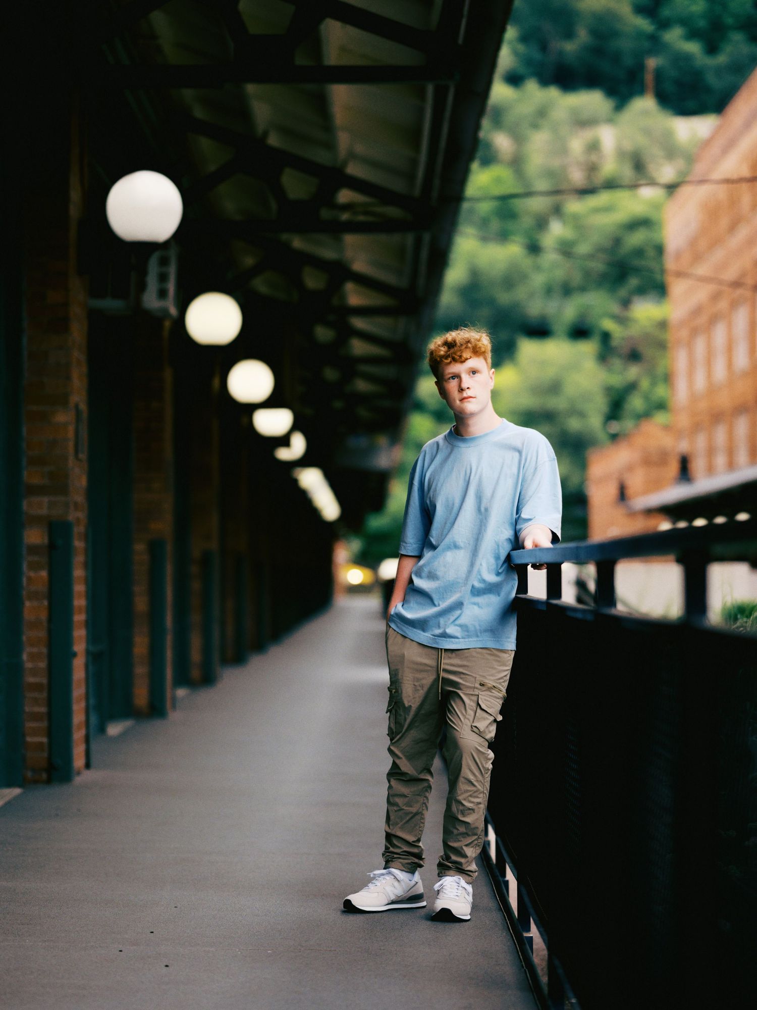 Young man in blue shirt and cargo pants poses on covered walkway with globe lights and green hillside backdrop.