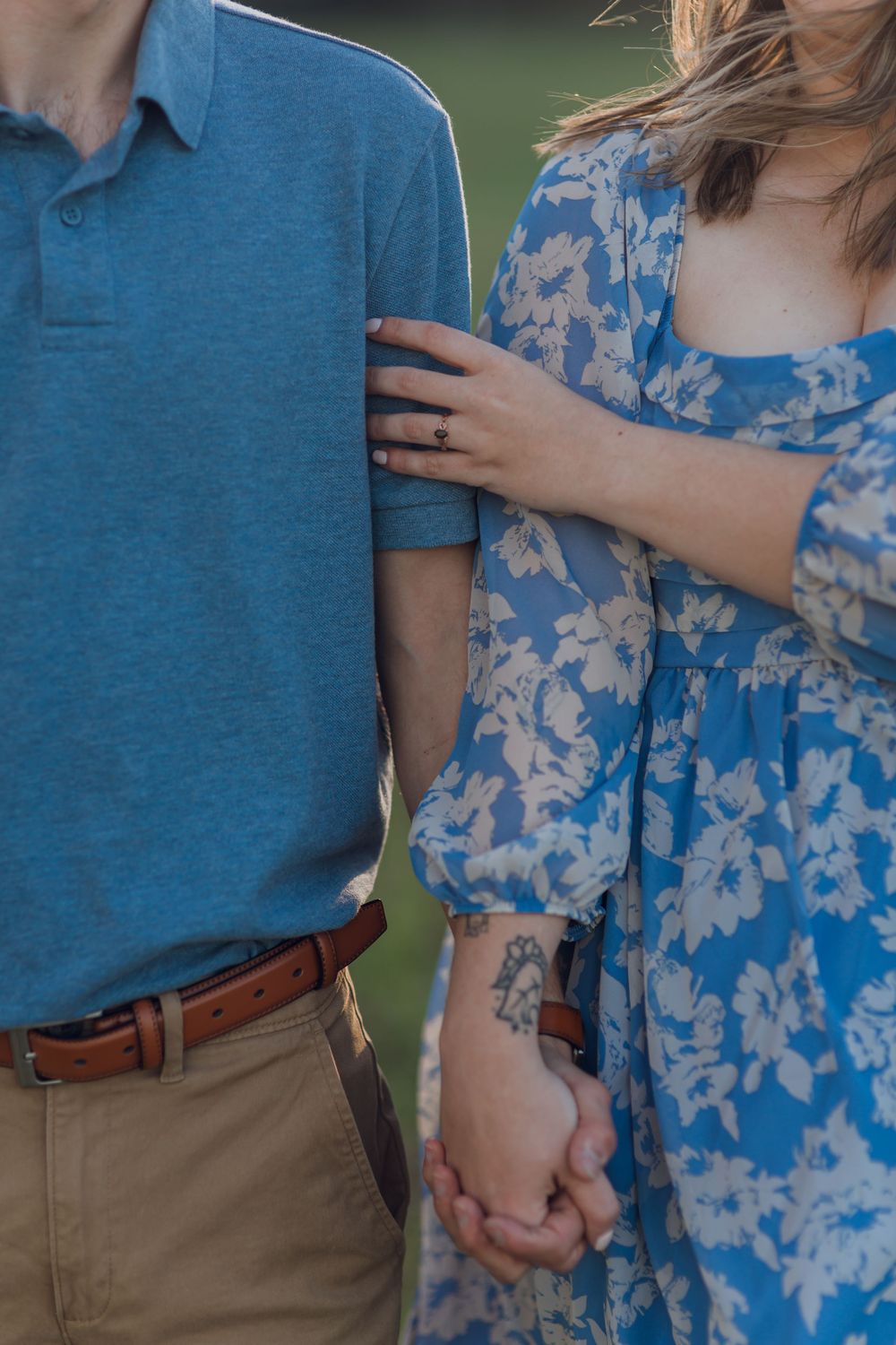 Couple holding hands, woman in blue floral dress with tattoo, man in teal polo and khaki pants, engagement ring visible.
