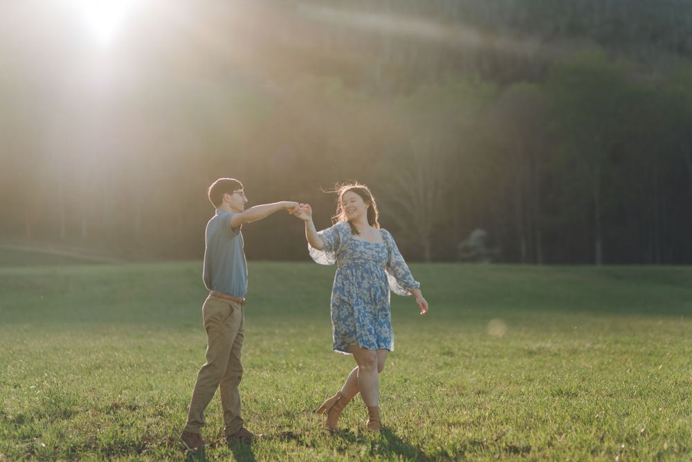Couple dancing together in a sunlit green field with golden backlight and trees in the background.