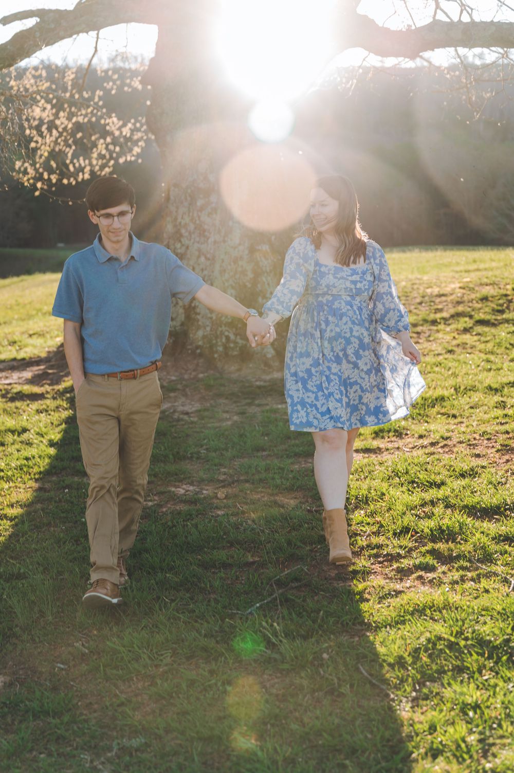 A couple holds hands walking outdoors in golden sunlight near a large tree on a grassy lawn.
