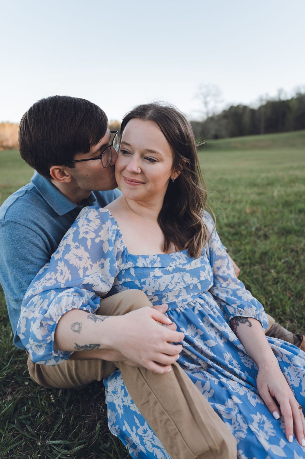A couple sits on green grass at sunset, man kissing woman's cheek as she smiles in a blue floral dress.