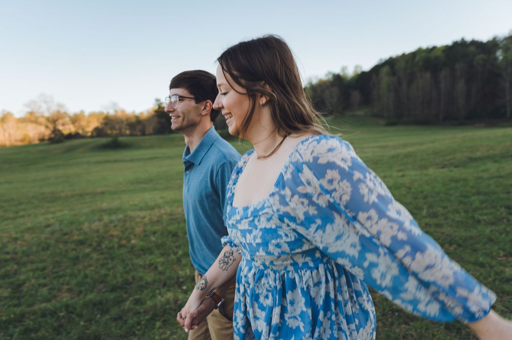 A couple walks hand in hand through a lush green field at sunset, smiling joyfully in casual outdoor attire.