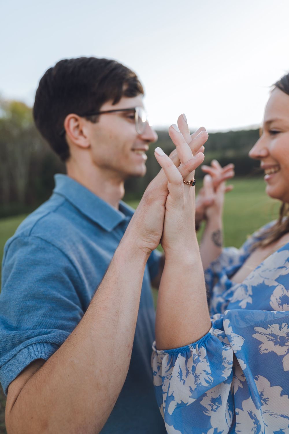A smiling couple holds hands outdoors, the woman wearing a floral blue dress and the man in a blue shirt.