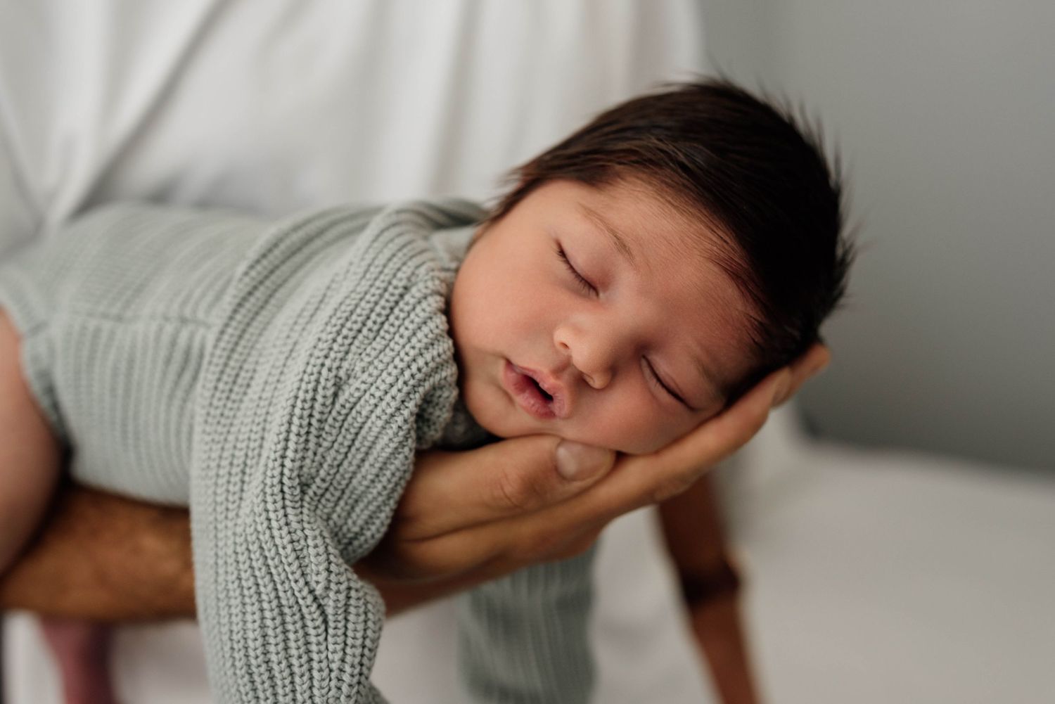 Sleeping newborn baby wrapped in sage green knit swaddle, cradled in adult hands on a wooden prop.