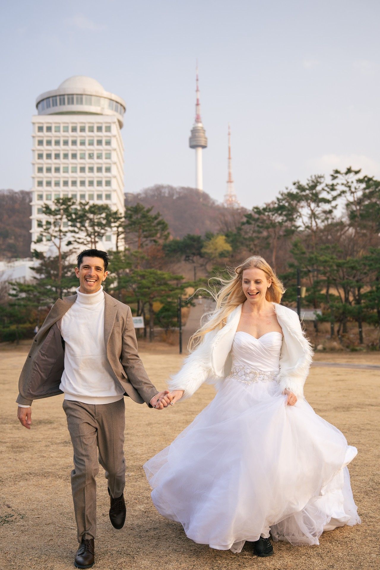 A couple in wedding attire holds hands and runs together with Seoul Tower and city buildings in the background.