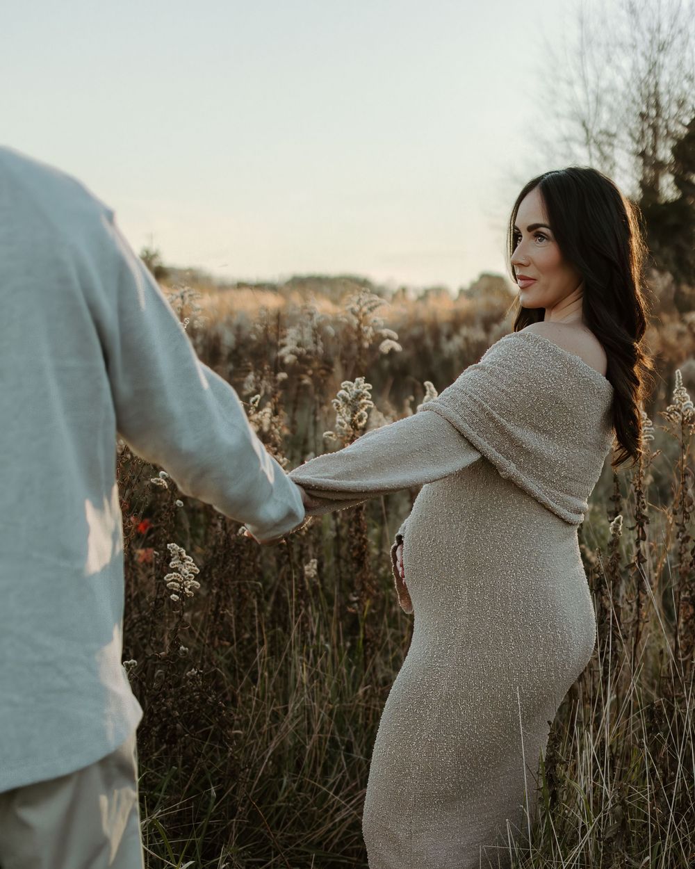 A couple holds hands in a rustic field setting during sunset while wearing cozy sweaters.