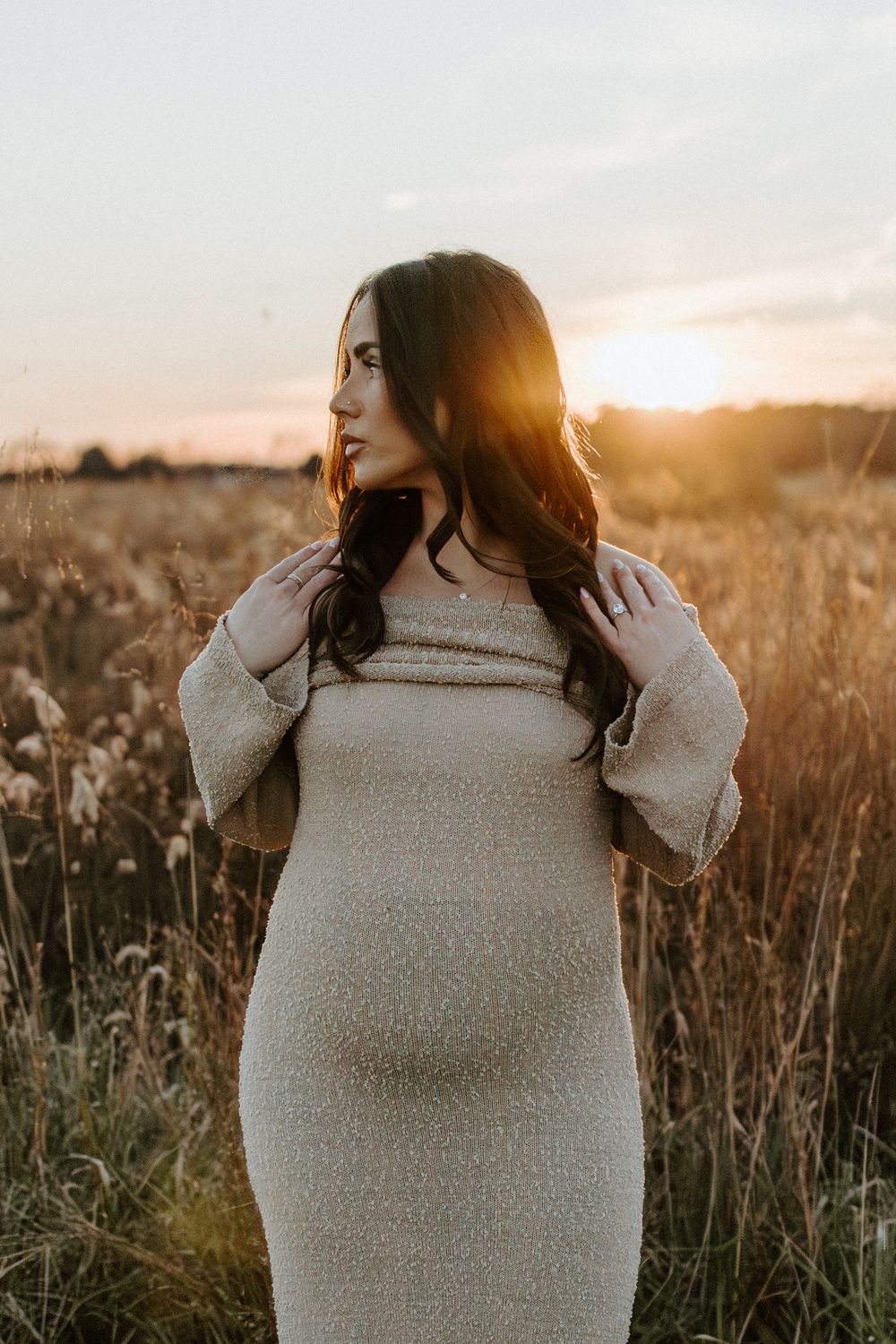 Woman in beige sweater dress poses in golden sunlit field during sunset.