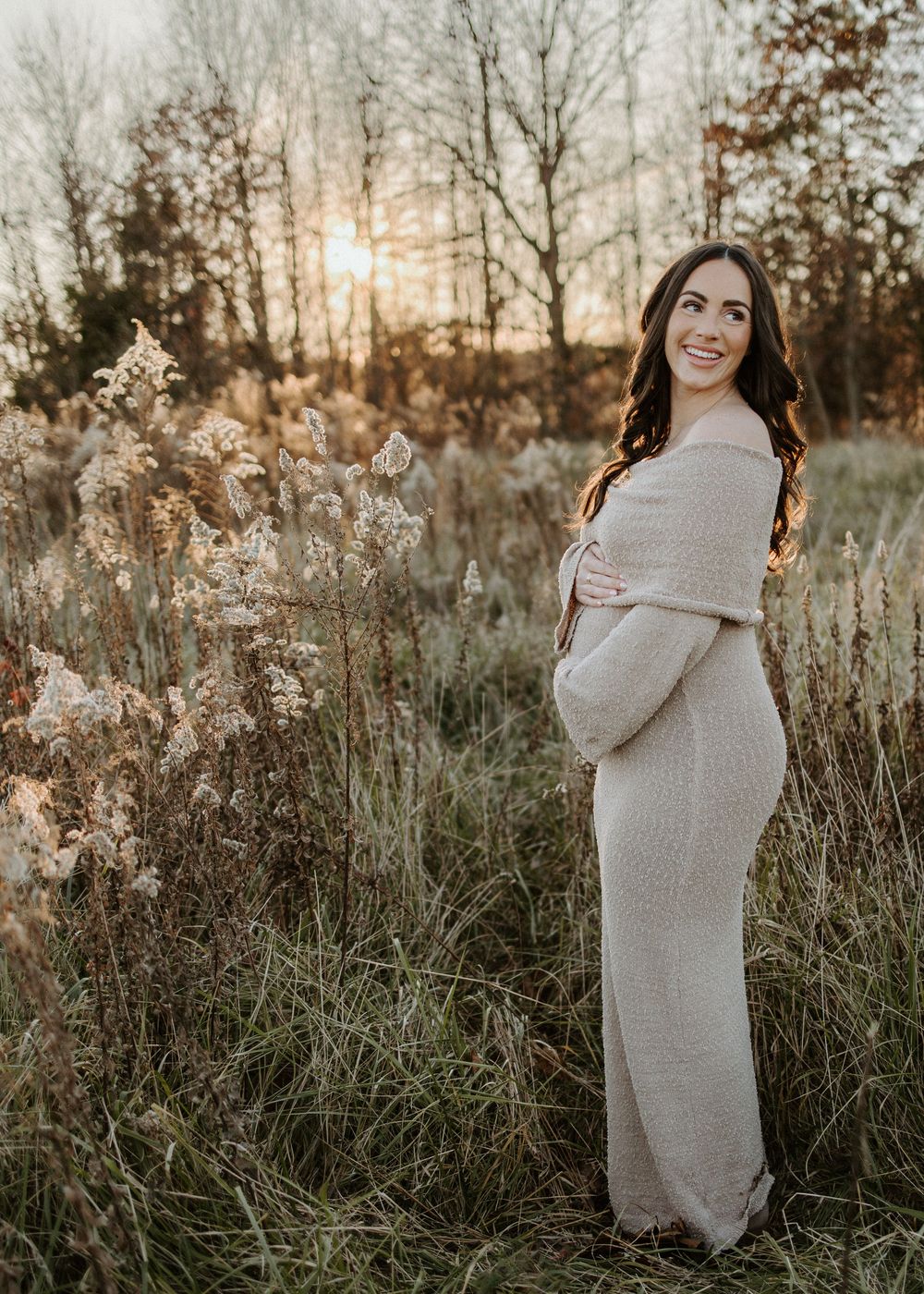 Maternity photo of woman in cream dress standing in golden field at sunset.