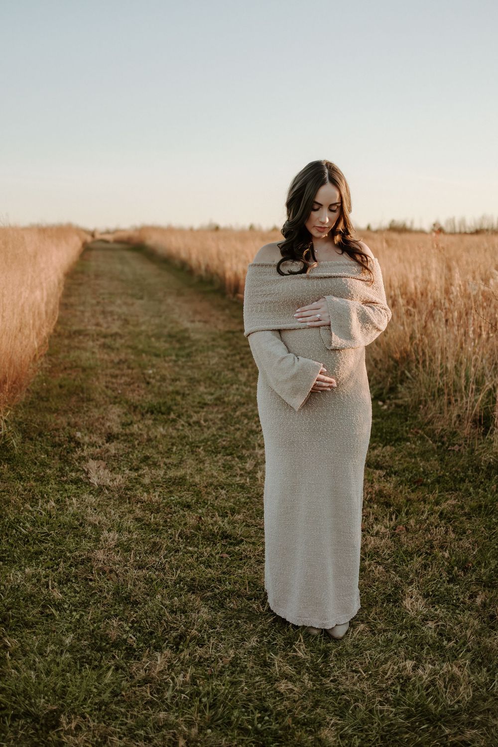 Woman in flowing beige dress stands on path through autumn field at sunset.