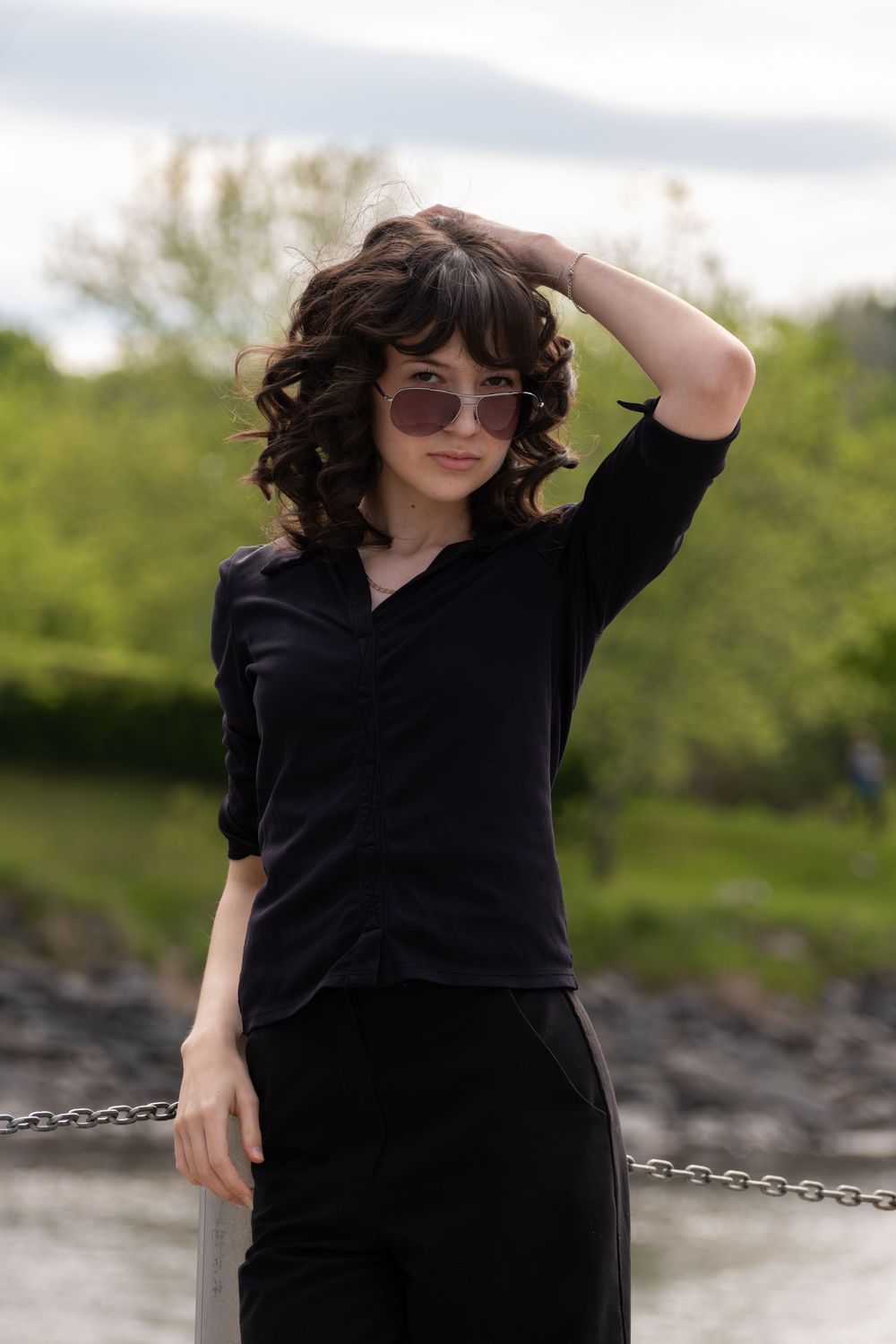 Young woman with curly hair and sunglasses posing outdoors in a black outfit near a chain fence with green trees.