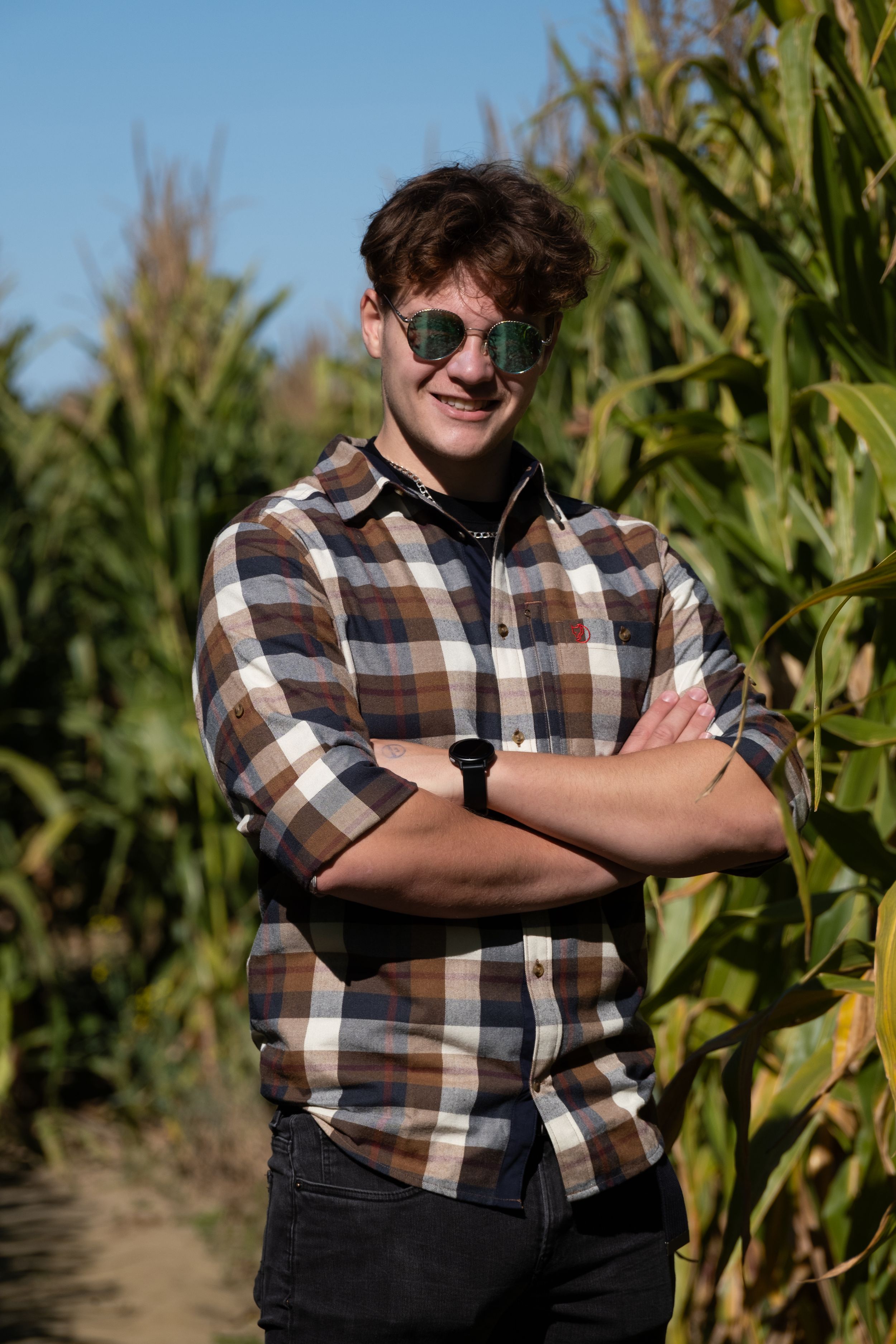 Figure in plaid shirt and sunglasses stands confidently in front of tall corn stalks.