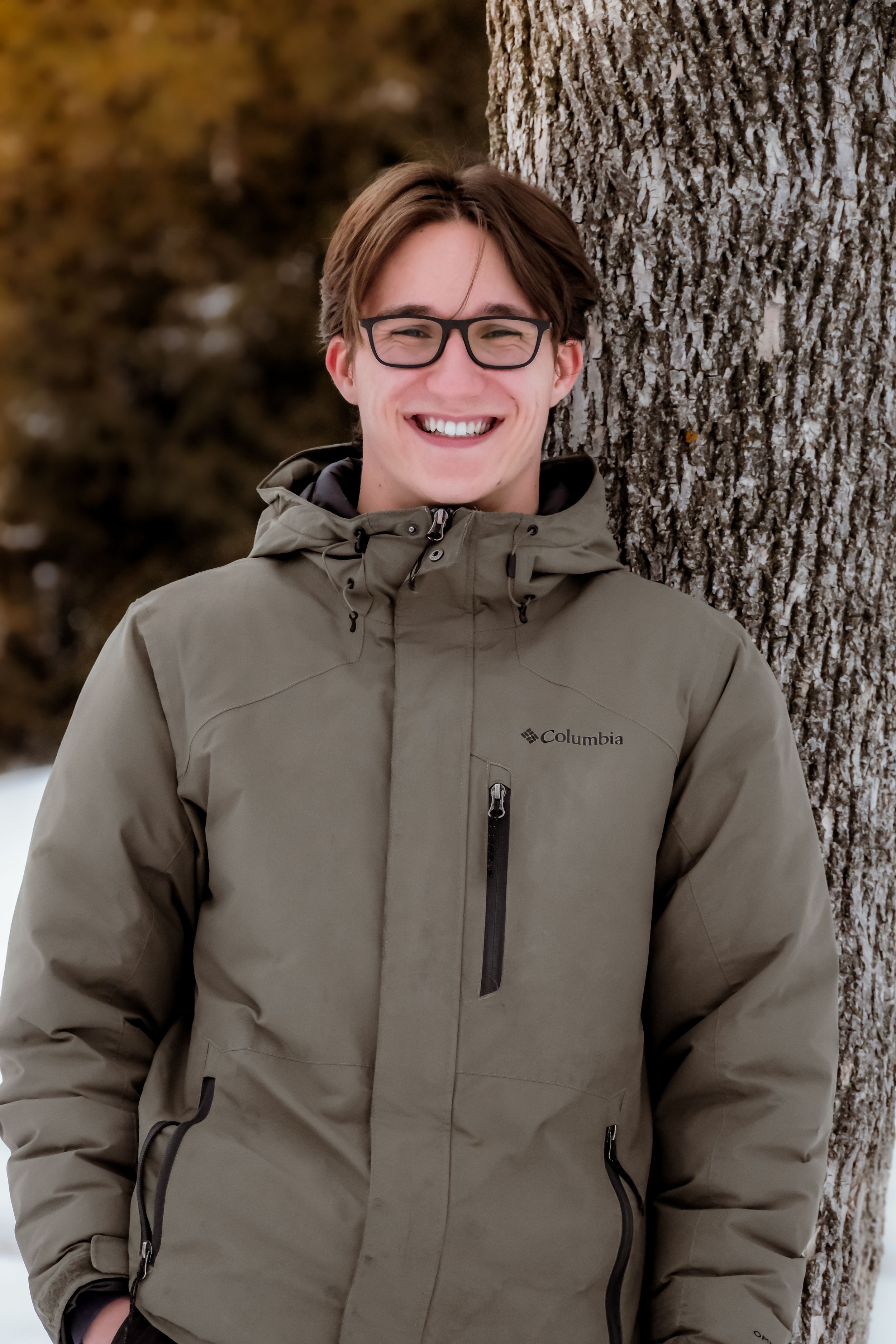 Person wearing a Columbia winter coat and glasses stands against a tree trunk in an outdoor setting.