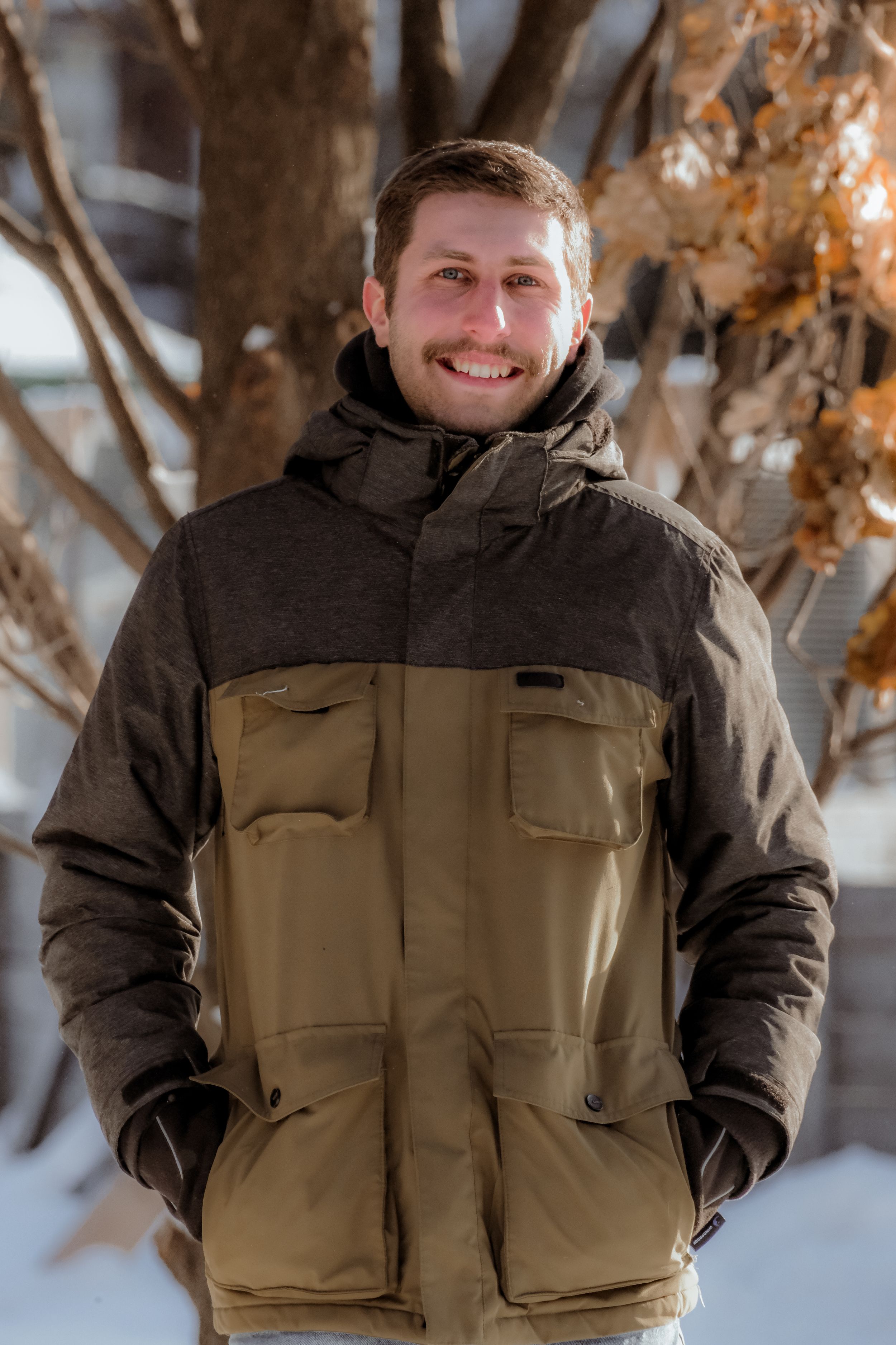 Person wearing a brown and black winter jacket smiles while standing in snowy outdoor setting with trees.
