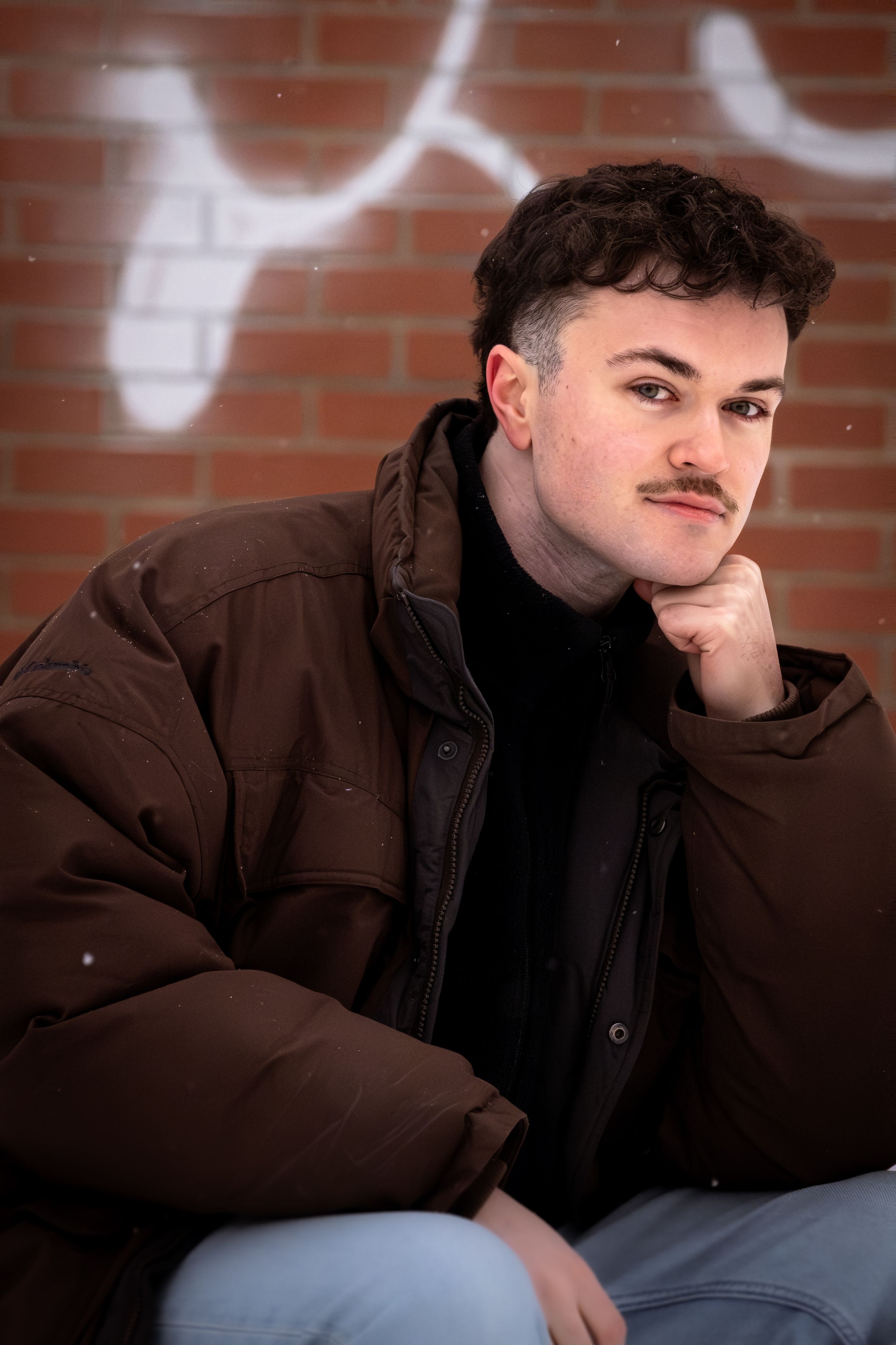 Person in brown jacket leans against brick wall in moody portrait series.