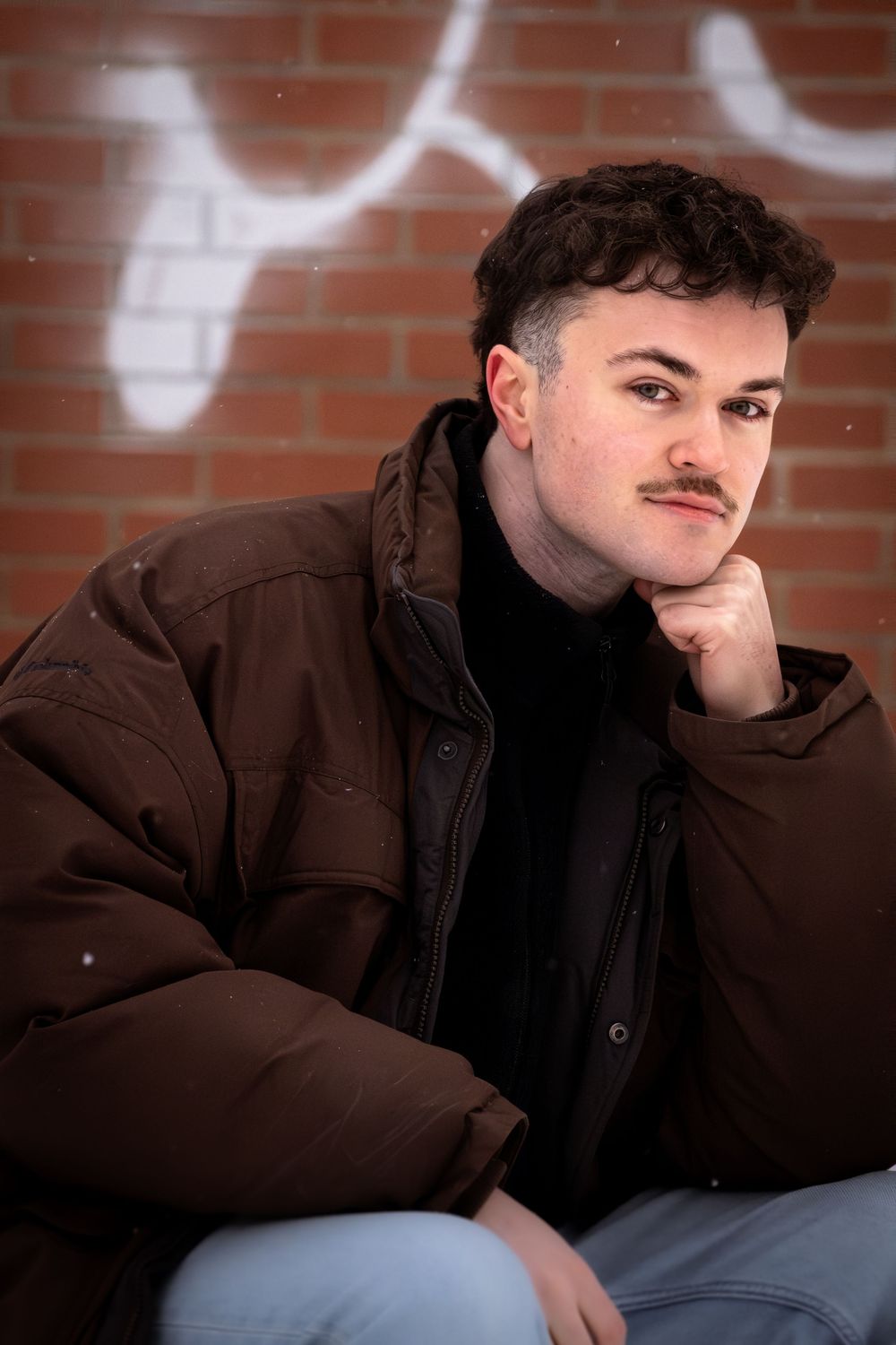 Person in brown jacket leans against brick wall in moody portrait series.