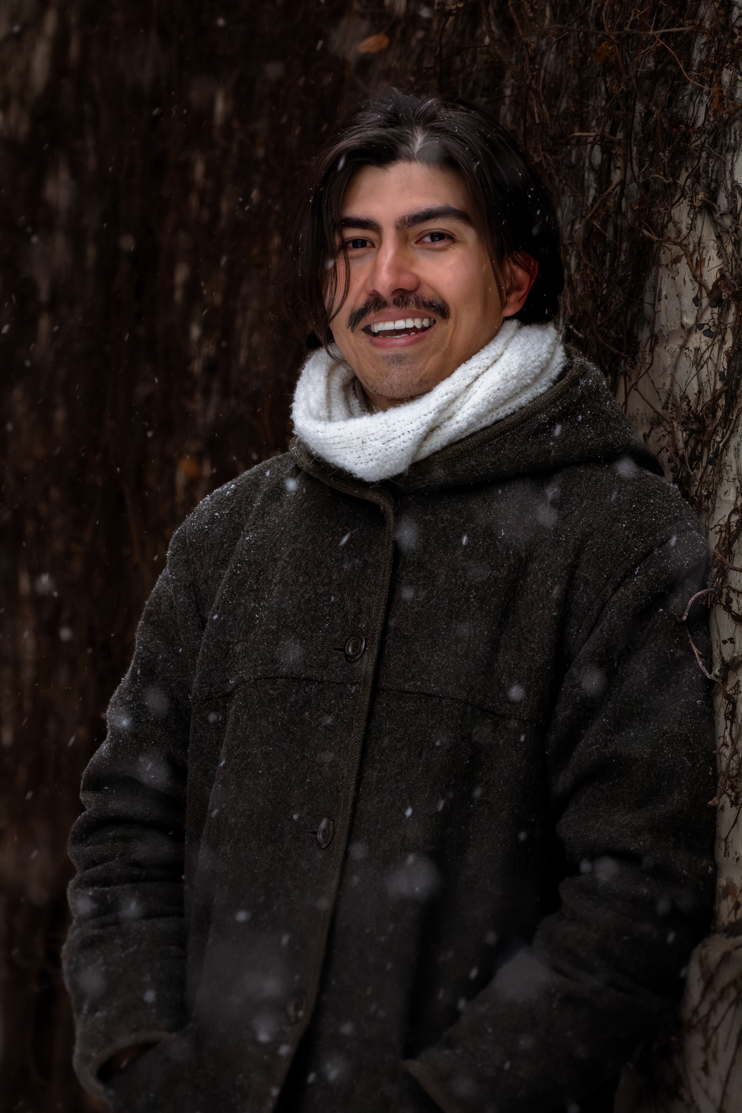 A person wearing a black winter coat and white scarf smiles while standing in front of snow-covered trees.