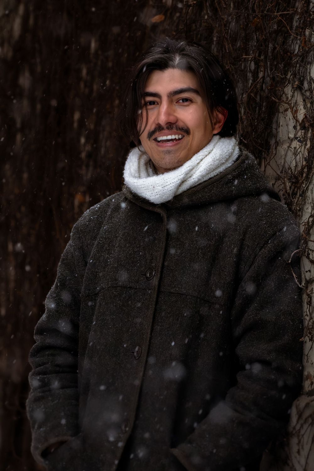 A person wearing a black winter coat and white scarf smiles while standing in front of snow-covered trees.