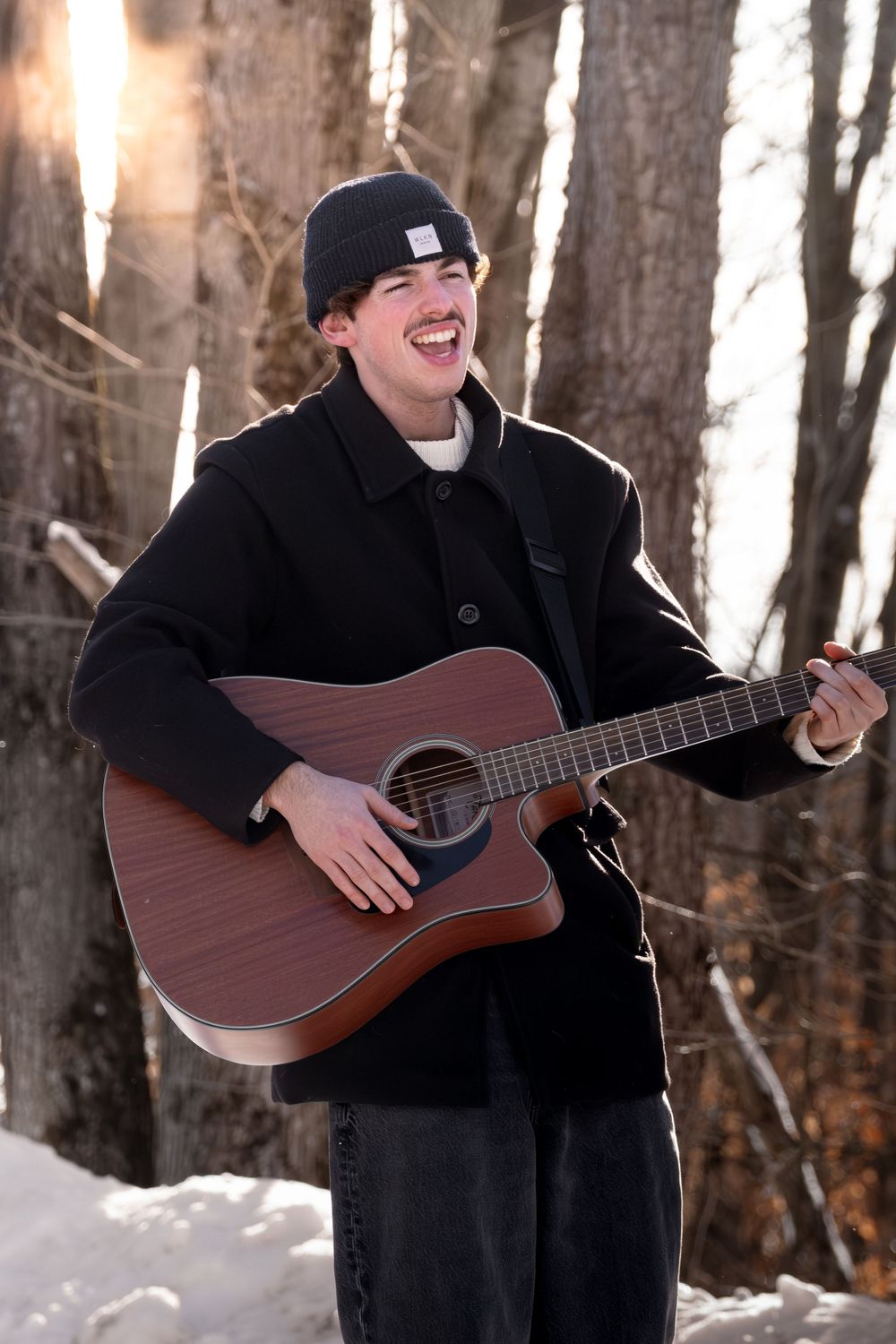 Person in black winter clothing plays acoustic guitar and laughs while standing in snowy forest.