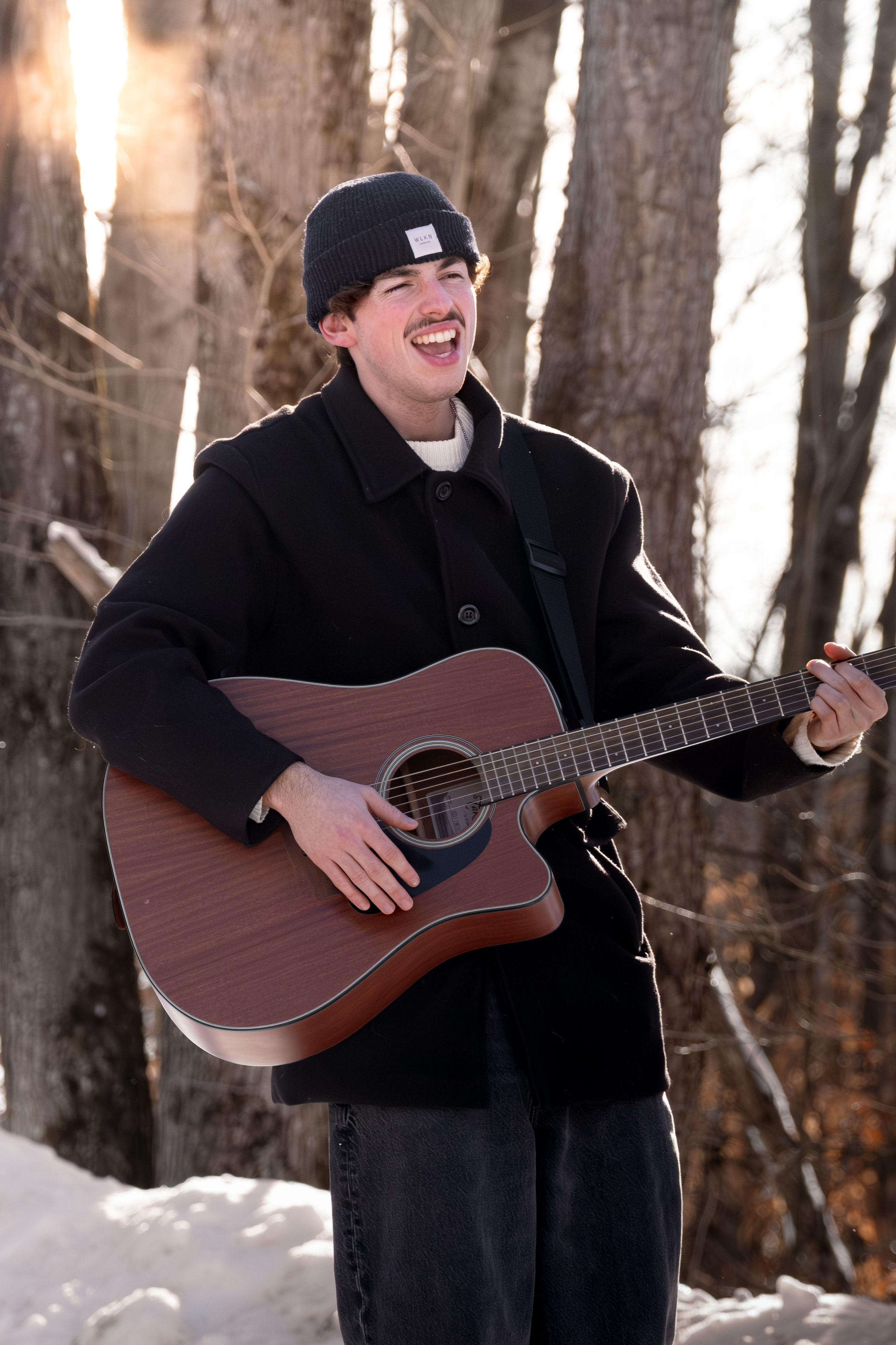 Person in black winter clothing plays acoustic guitar and laughs while standing in snowy forest.